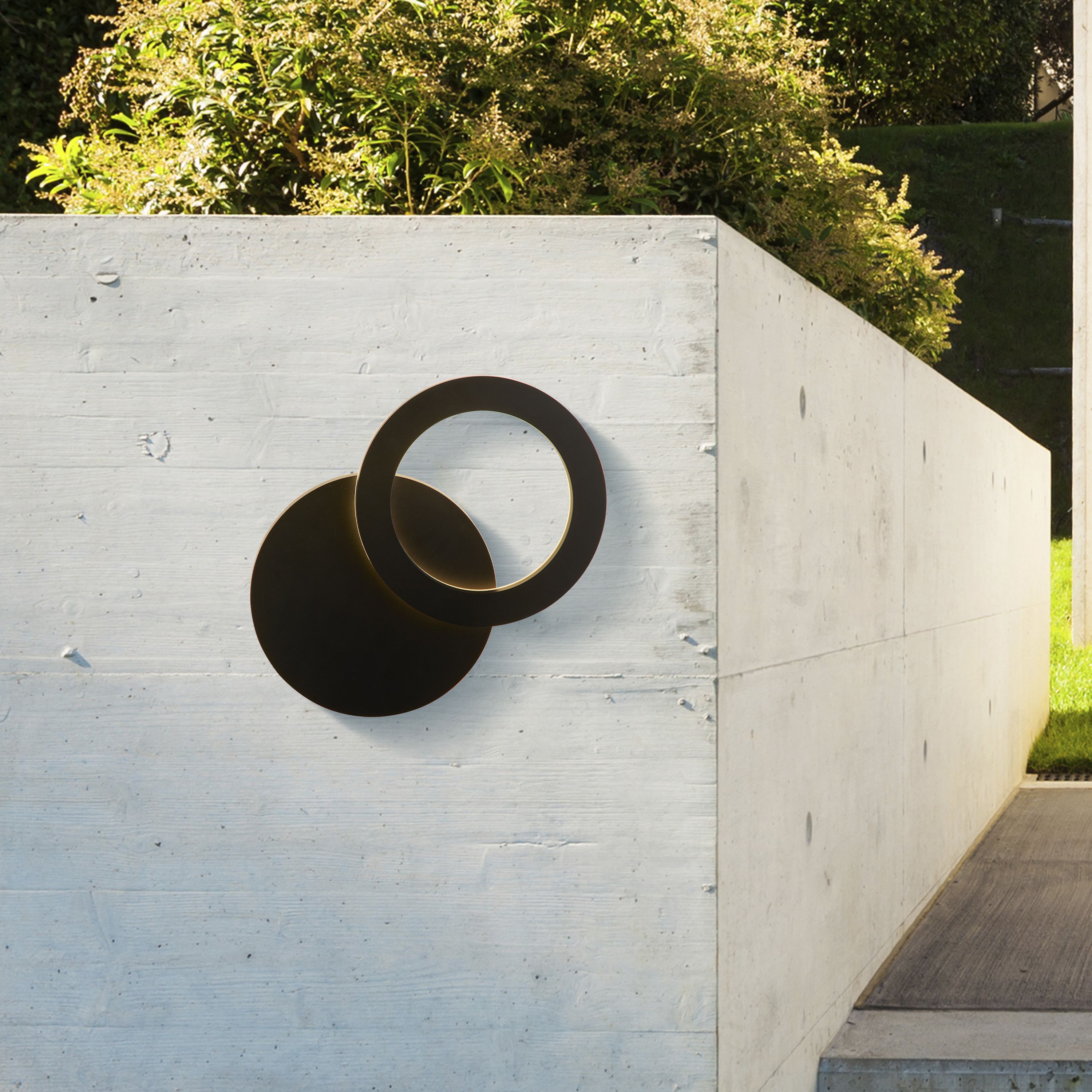 Searchlight Hyde LED outdoor wall light installed on a concrete garden wall in daylight, shown unlit beside greenery