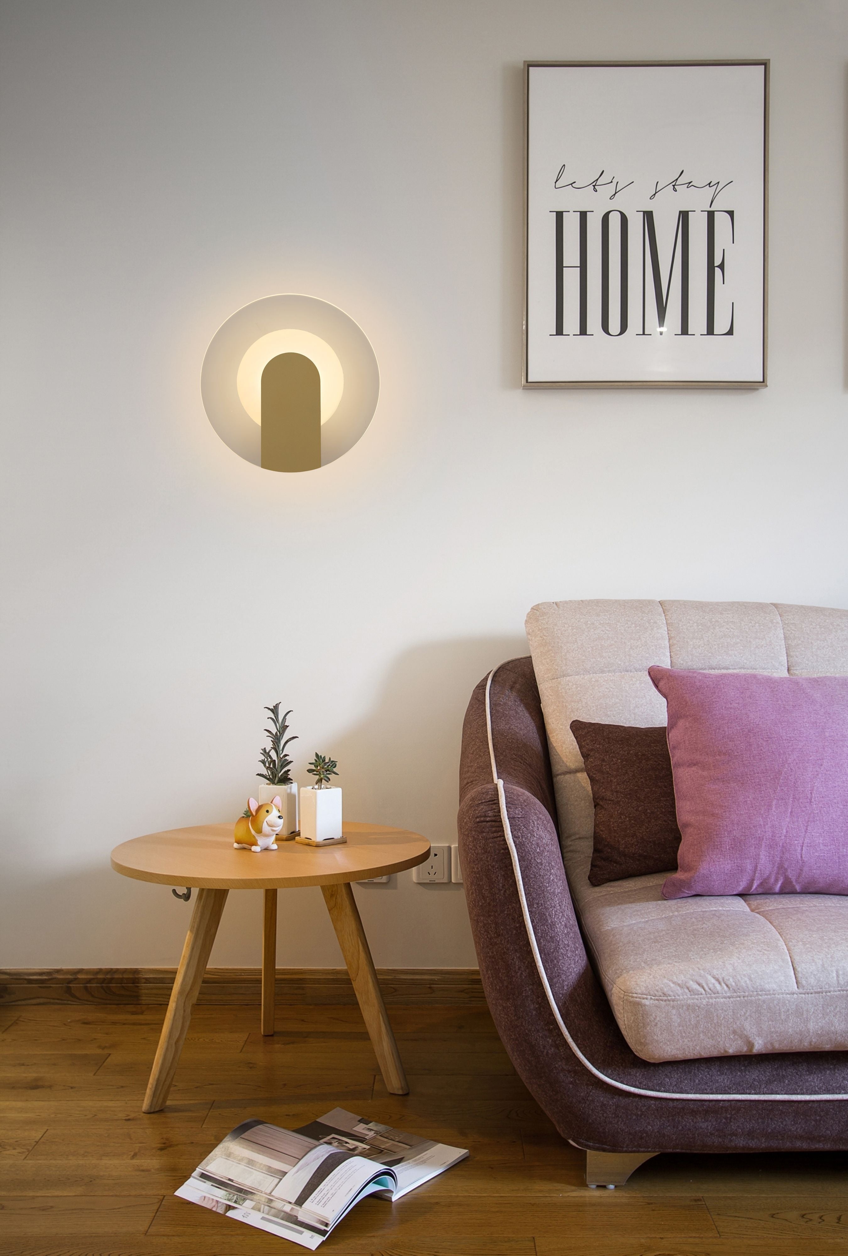 Target wall light lit and mounted above a side table with potted plants and a framed Let's Stay Home print, beside a sofa with pink and brown cushions