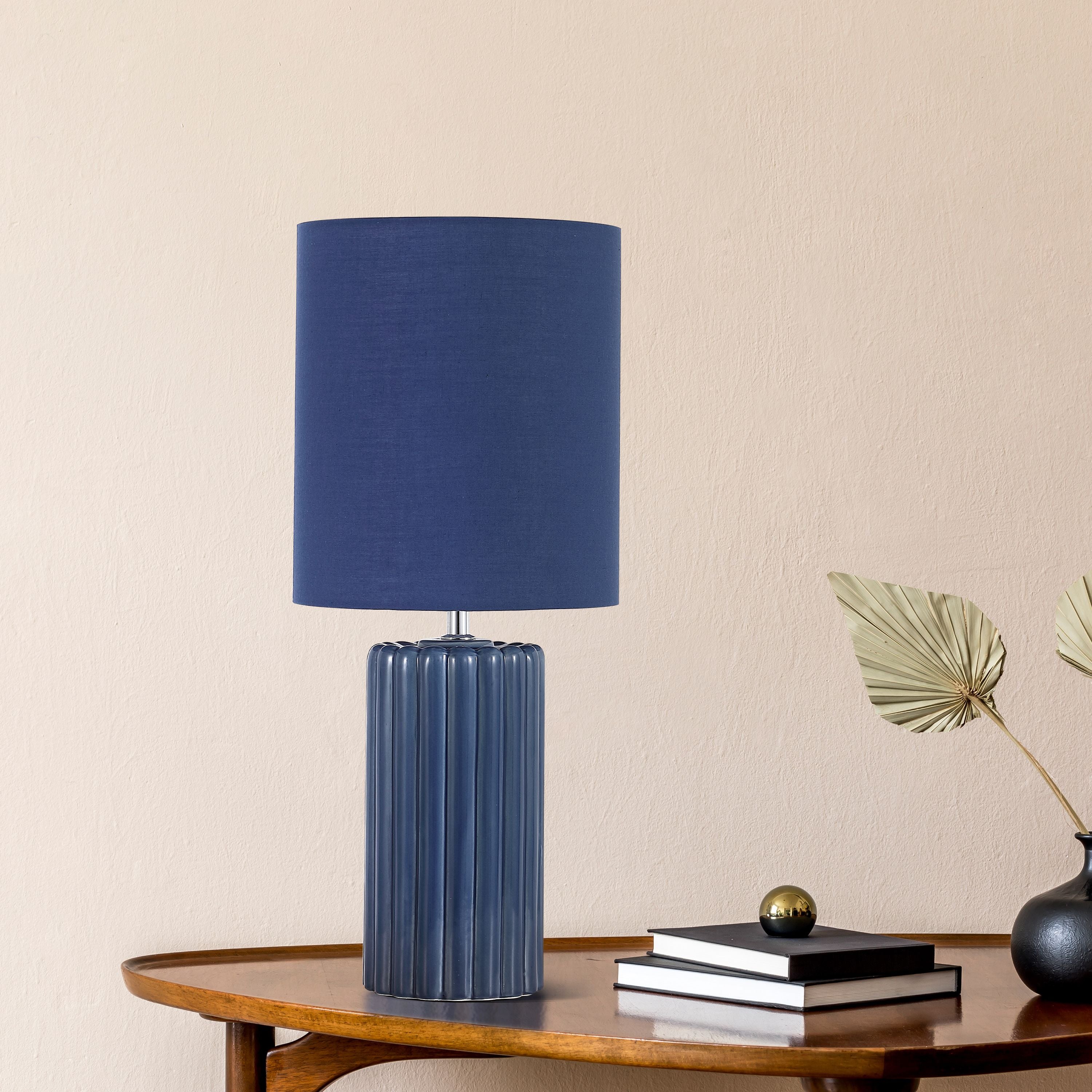 Groove navy blue table lamp styled on a wooden console table beside a stack of books, a small brass ornament and a dried palm frond