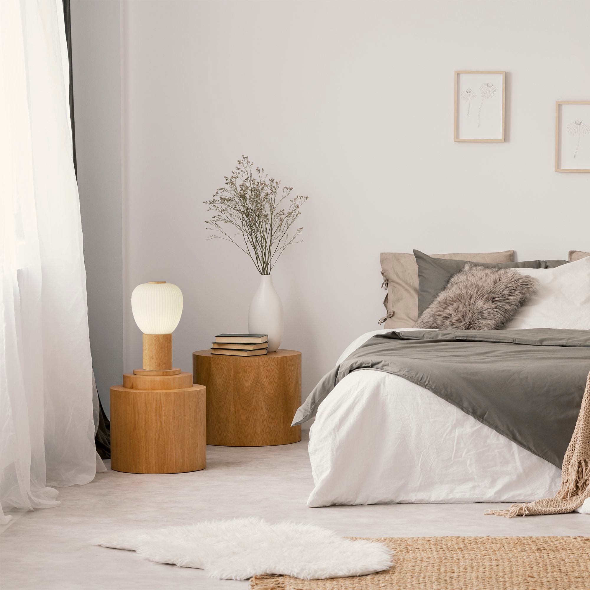 Searchlight Ella table lamp switched on beside a bed in a styled bedroom, standing on a round wooden side table next to a vase and stacked books