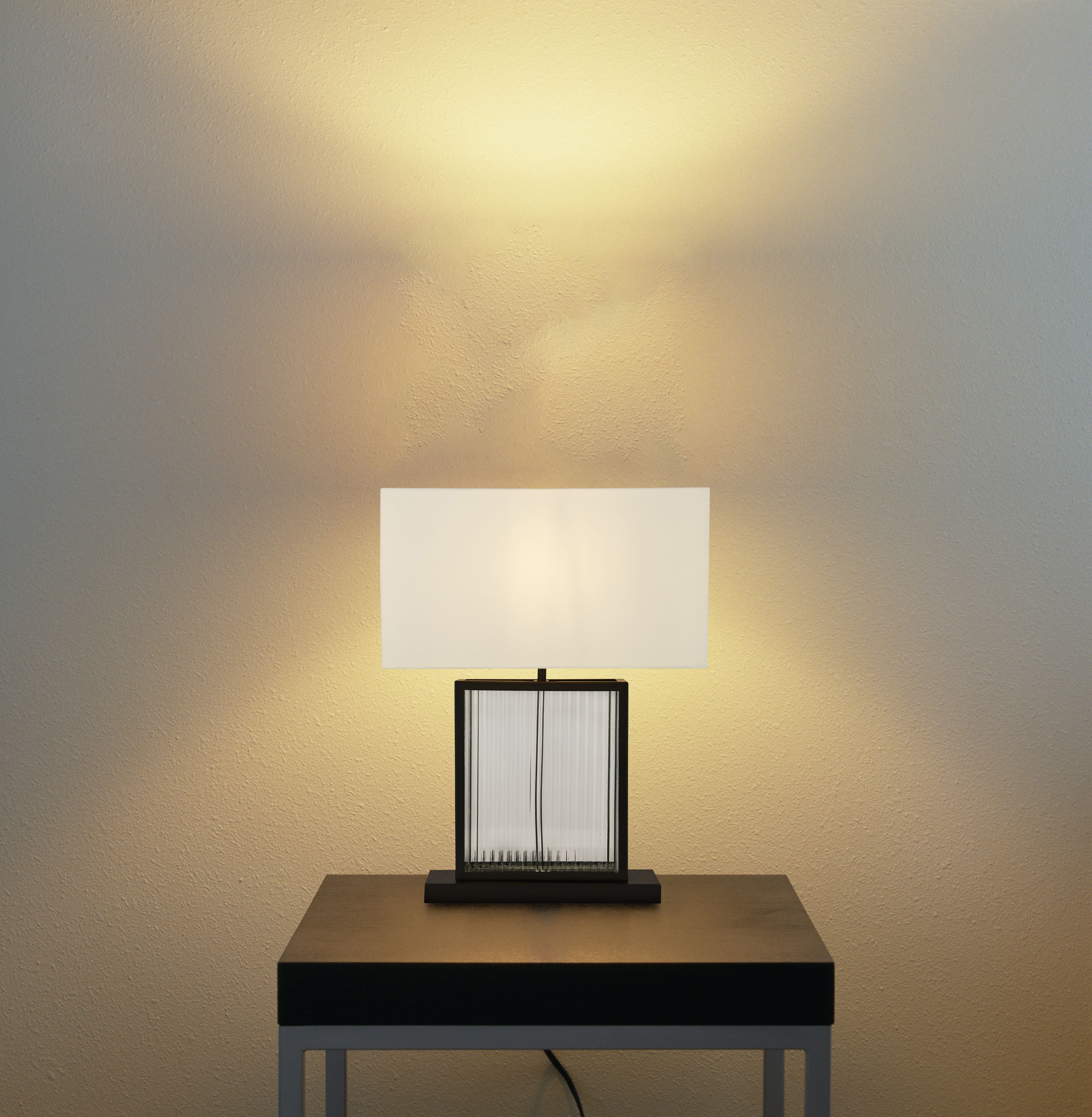 Clarendon table lamp switched on and glowing warmly on a wooden side table against a plain wall