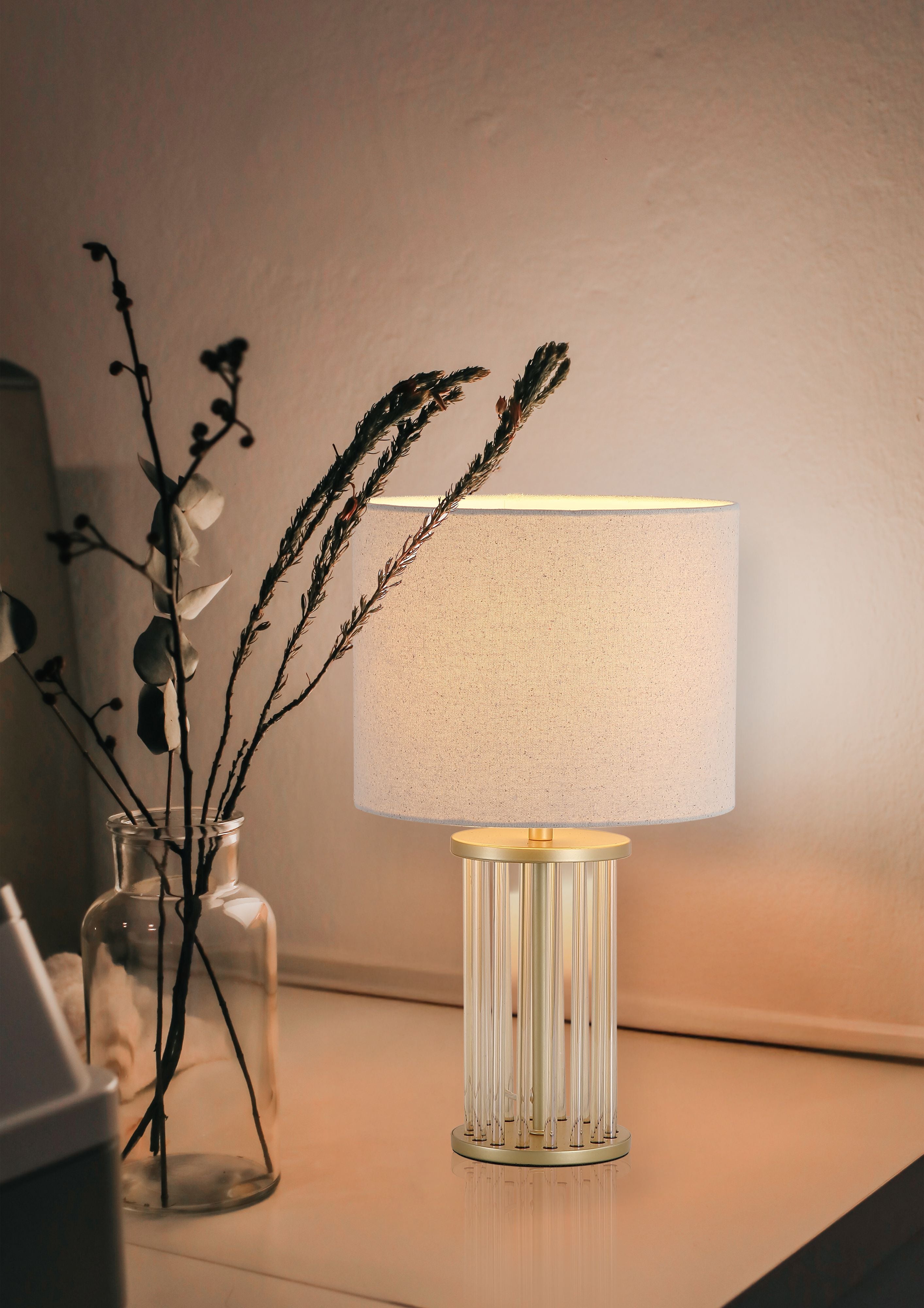 Swizzle table lamp in gold styled on a bedside table next to a glass vase of dried flowers, lamp switched on with a warm glow