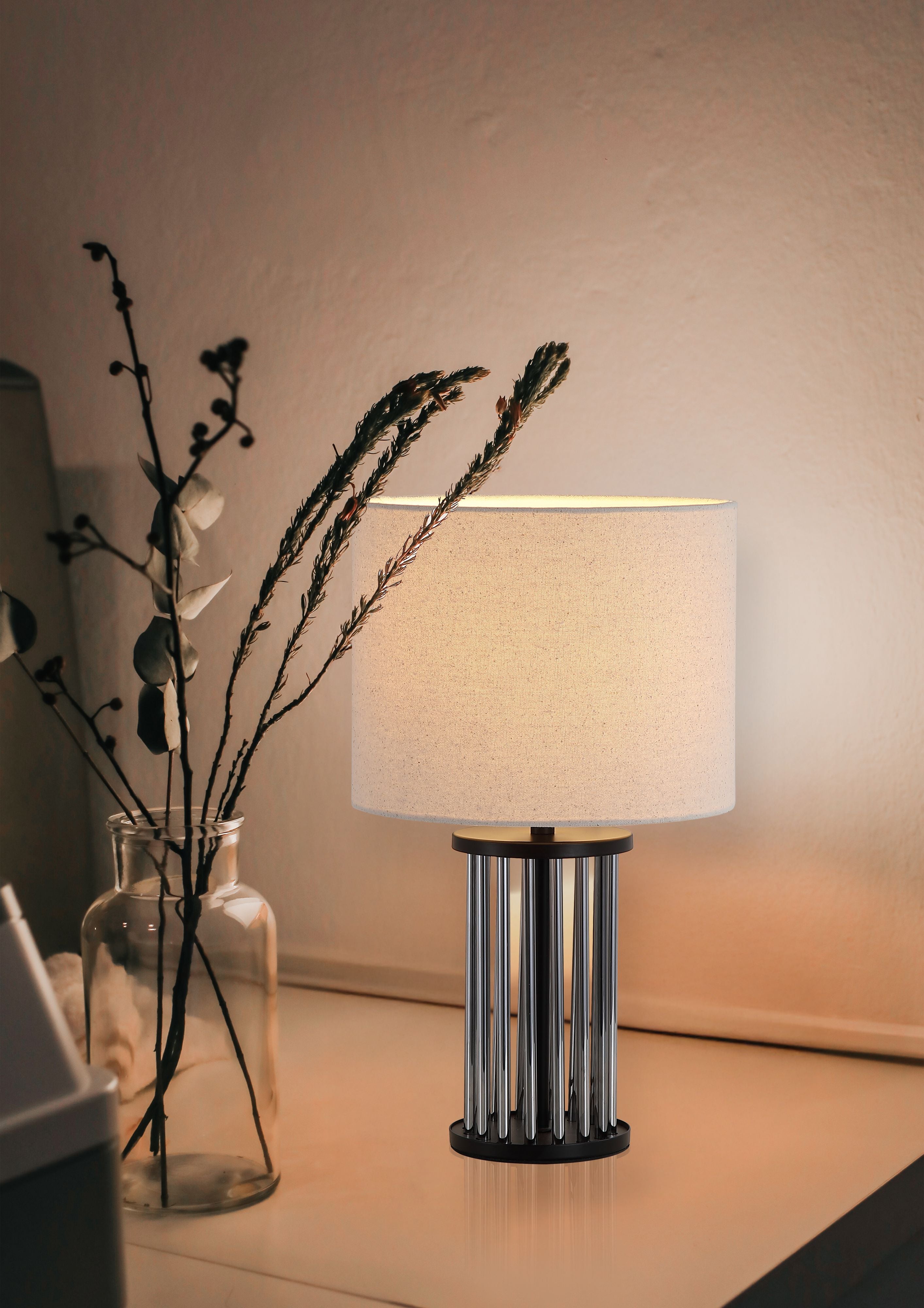 Swizzle table lamp in matt black styled on a bedside table next to a glass vase of dried flowers, lamp switched on with a warm glow