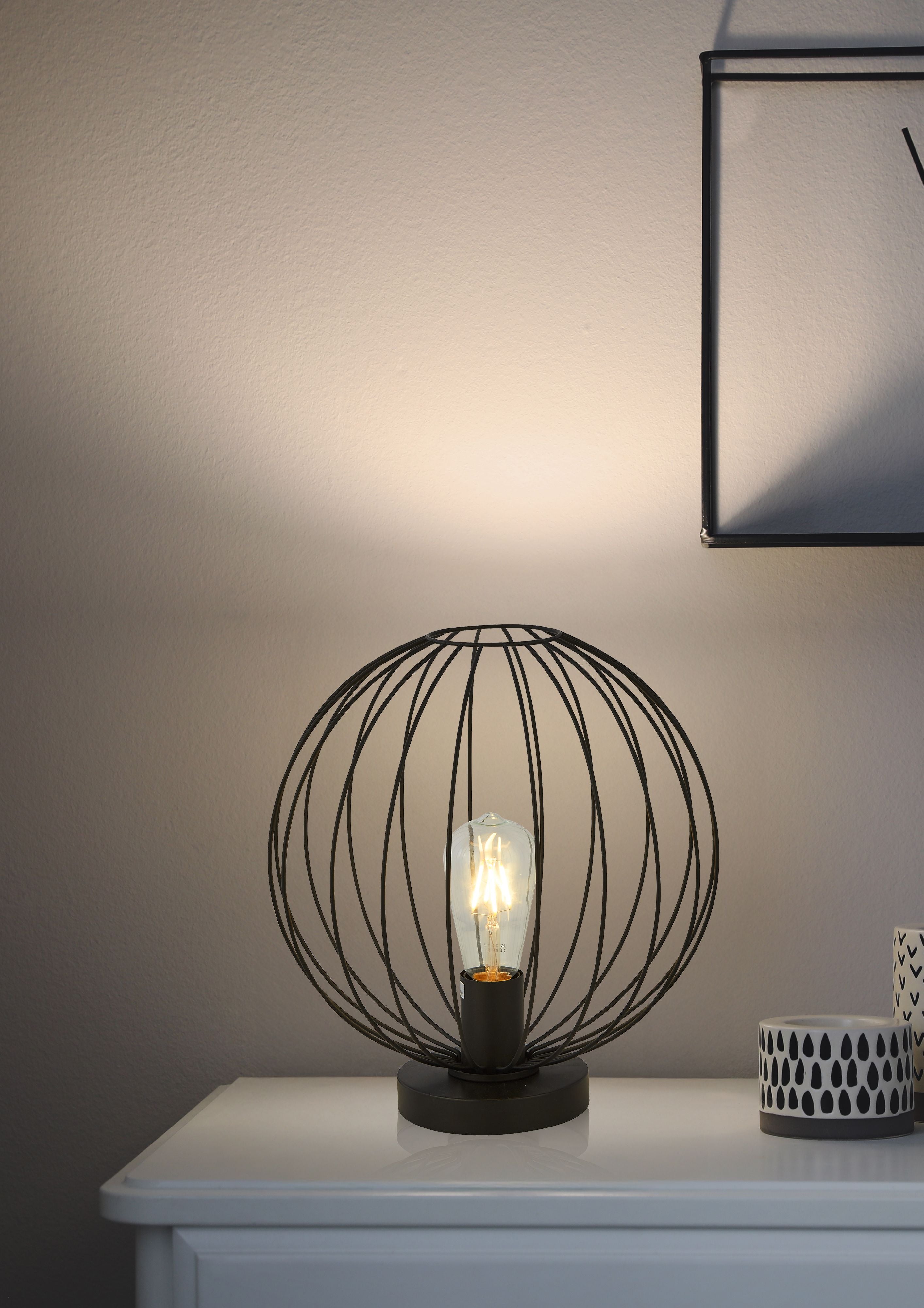 Searchlight Rondo table lamp styled on a white bedside table beneath a framed mirror, wire cage shade glowing warmly next to patterned ceramic pots