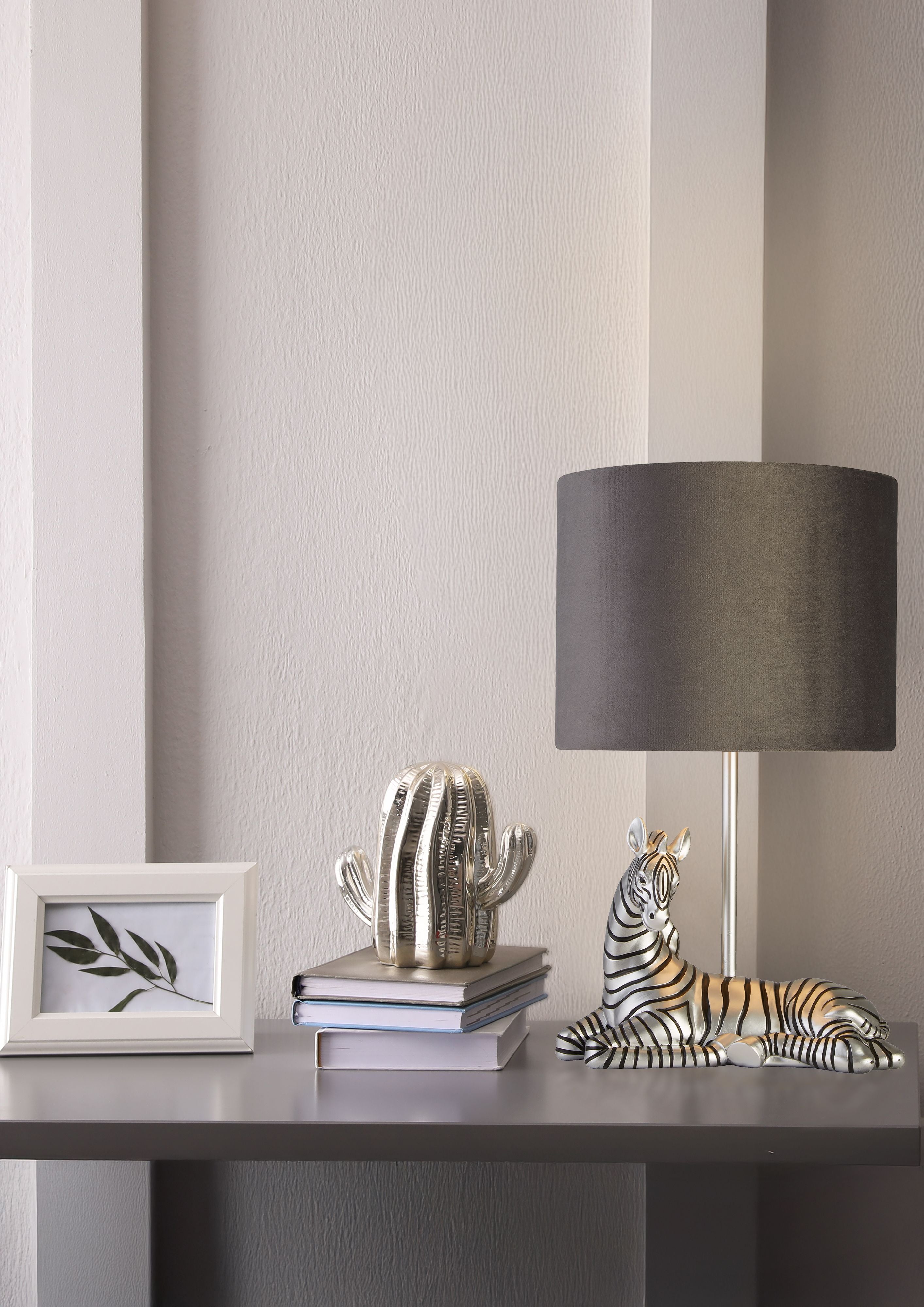 Searchlight Zebra table lamp with charcoal velvet shade styled on a grey console table beside a silver cactus ornament, stacked books and a white framed picture