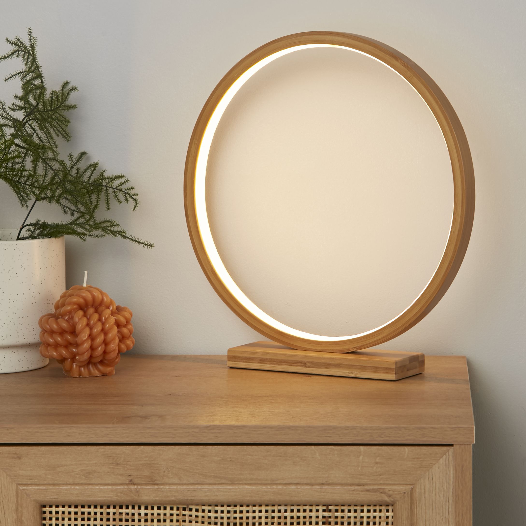 Searchlight Malibu LED table light glowing warm white on a wooden sideboard with a rattan front, beside a potted fern and a knotted orange candle