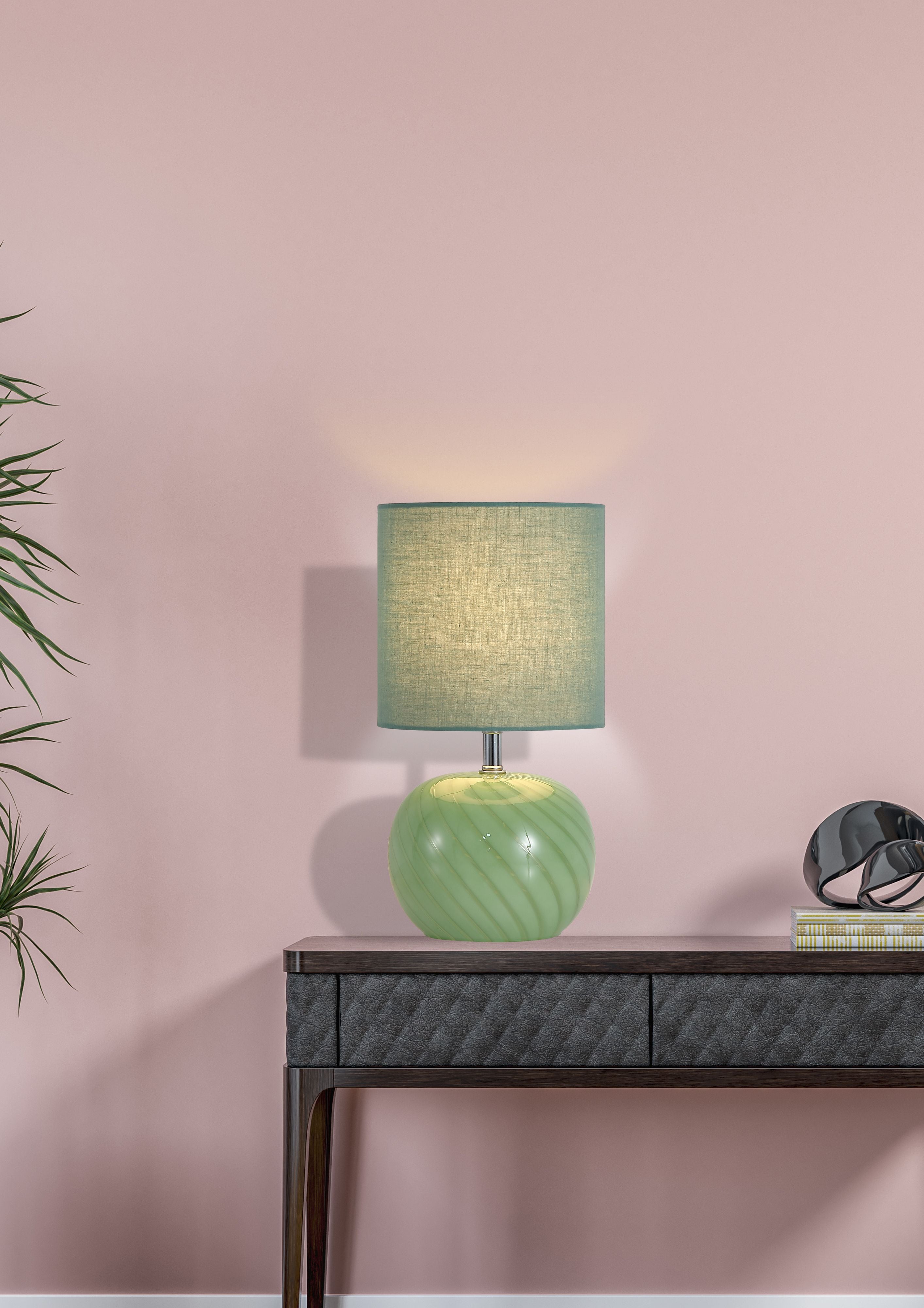 Fete green glass table lamp styled on a dark console table against a pink wall, lit with a warm glow, with a plant and decorative sphere nearby