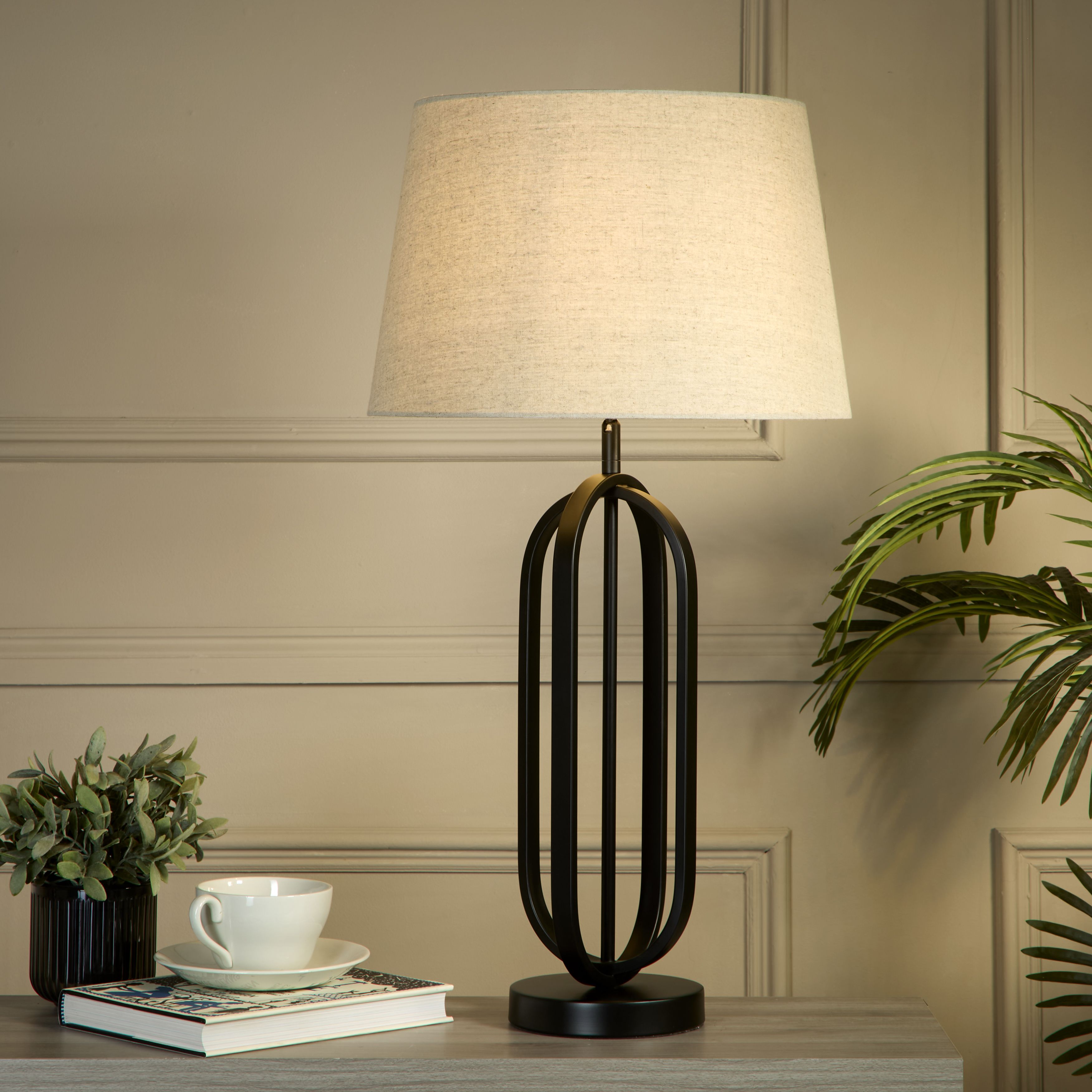 The lamp styled on a grey table against a taupe panelled wall, with a teacup and saucer, a book and potted plants either side, its natural linen shade glowing warm light