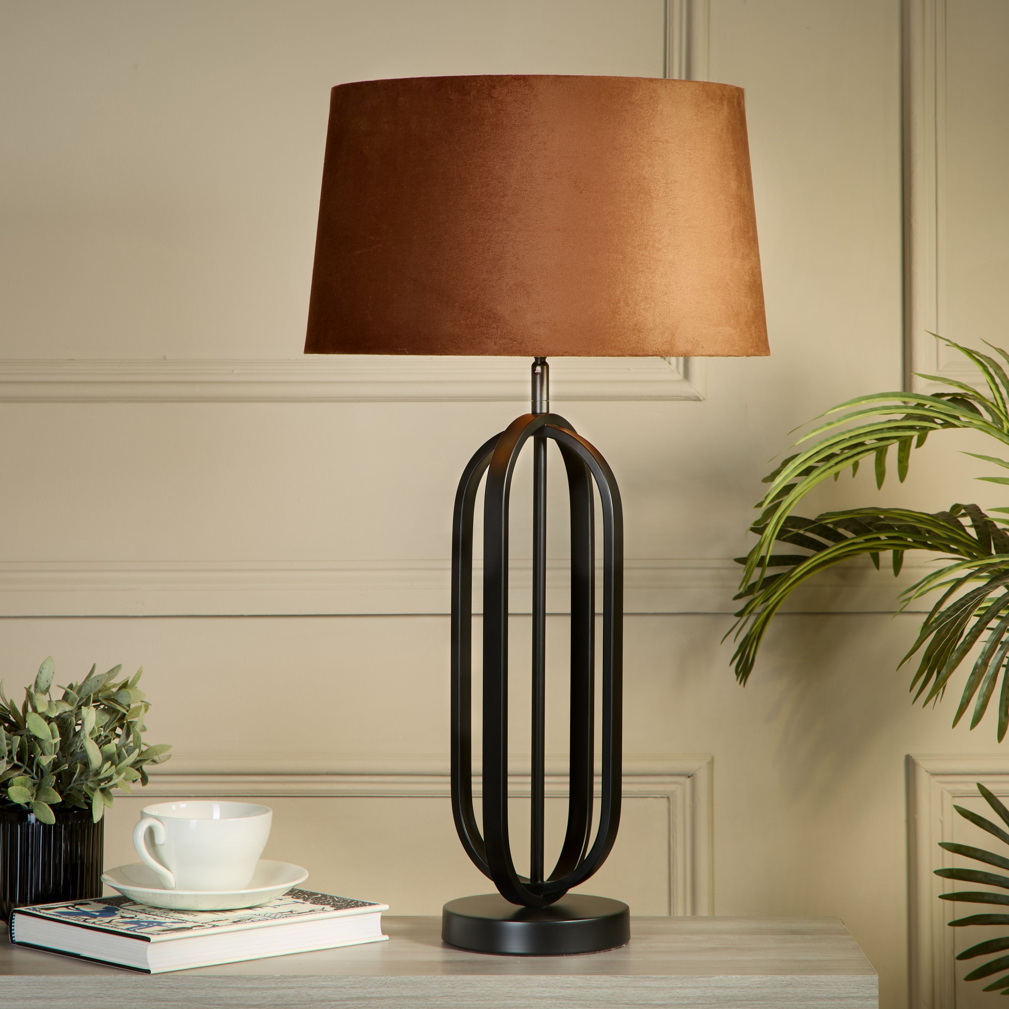The lamp styled on a grey table against a beige panelled wall, with a teacup and saucer, a book and potted plants either side, its burnt orange velvet shade glowing warm light
