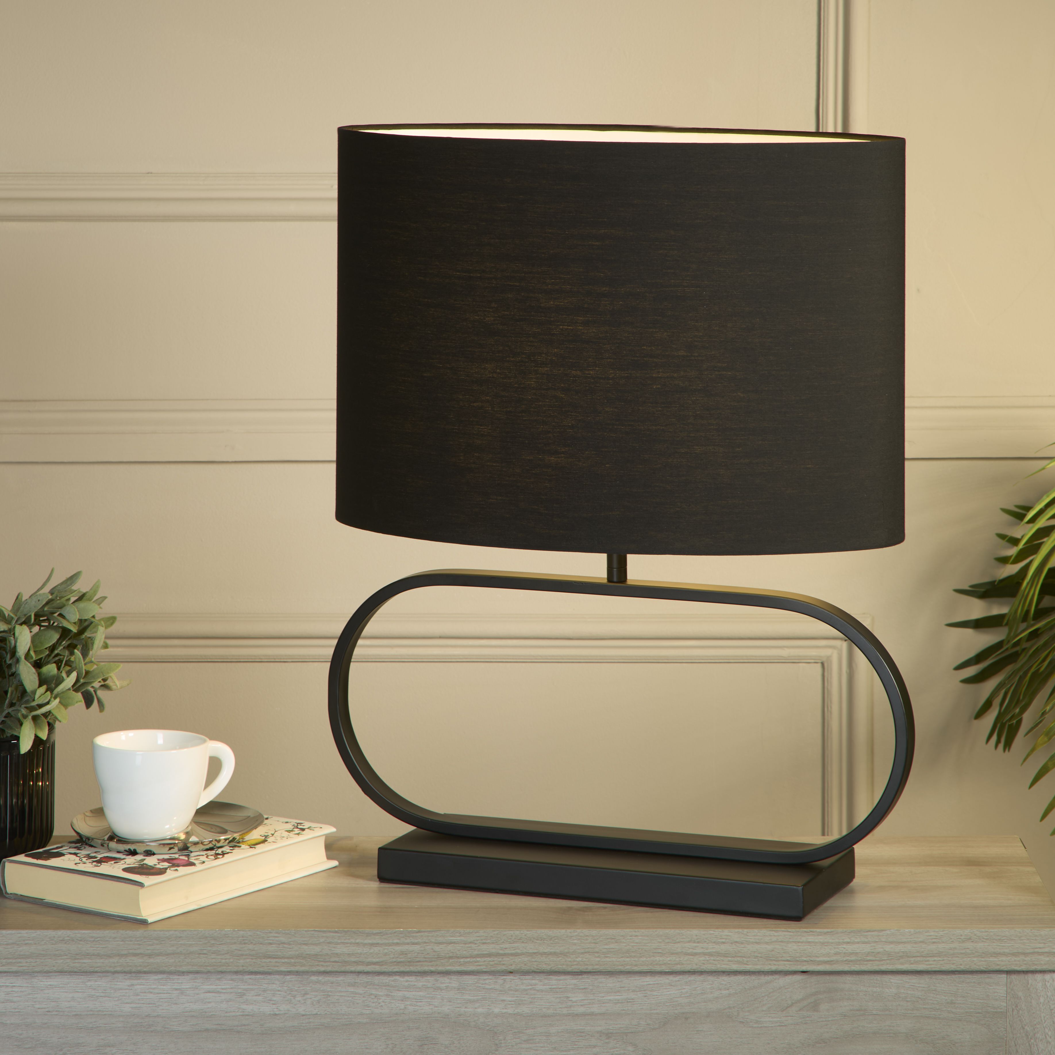 Sleek table lamp in black styled on a living room sideboard beside a book, a cup and saucer, and a small vase of foliage, shade glowing warm white.