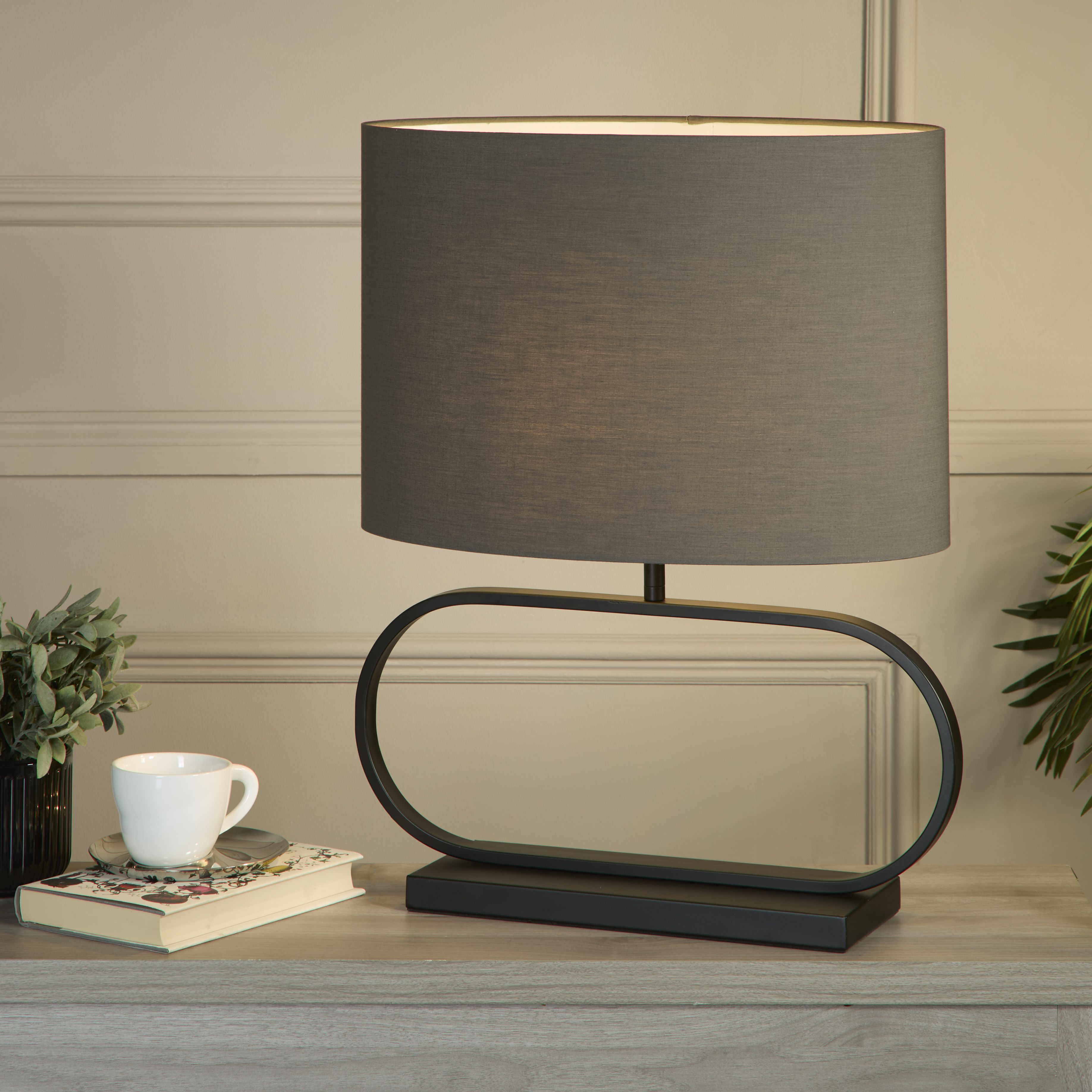 Sleek table lamp with a grey shade styled on a living room sideboard beside a book, a cup and saucer, and a small vase of foliage, shade glowing warm white.