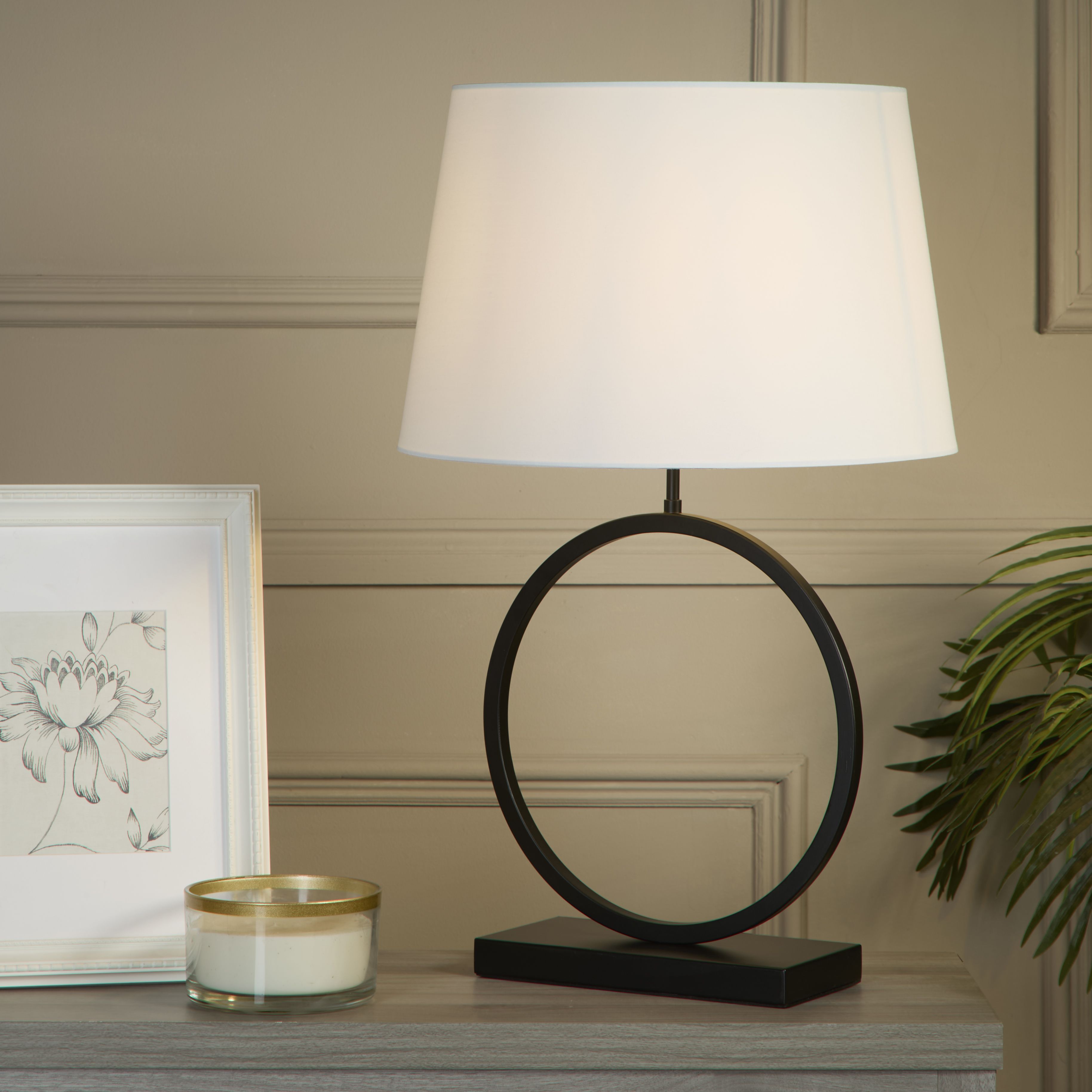 Elegant Table Lamp with white cotton shade lit up on a wooden bedside table next to a framed print, candle and a leafy plant