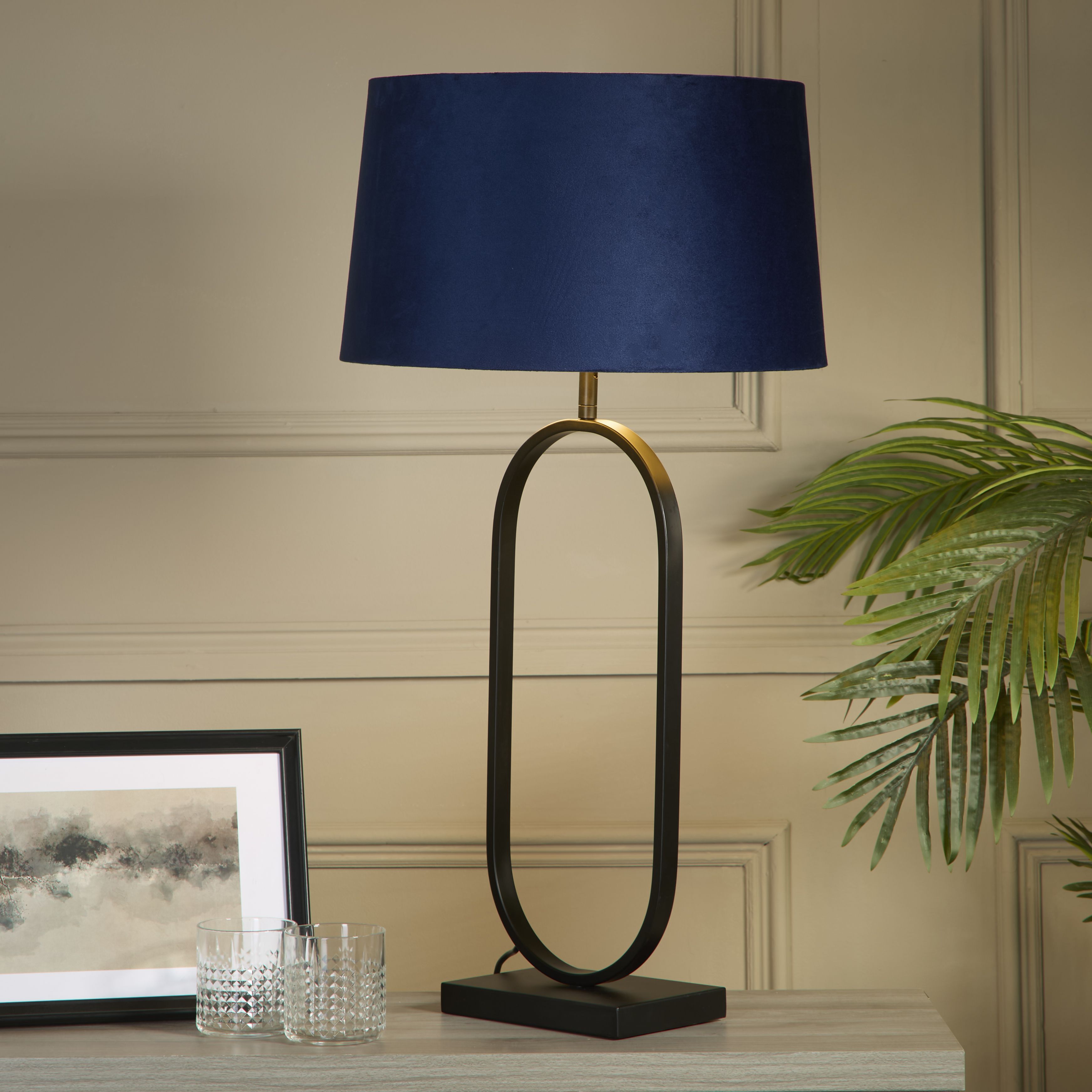 Modish table lamp in matt black with a navy velvet shade styled on a bedroom nightstand beside a picture frame, glass tumblers and a potted plant, shade glowing warm white.