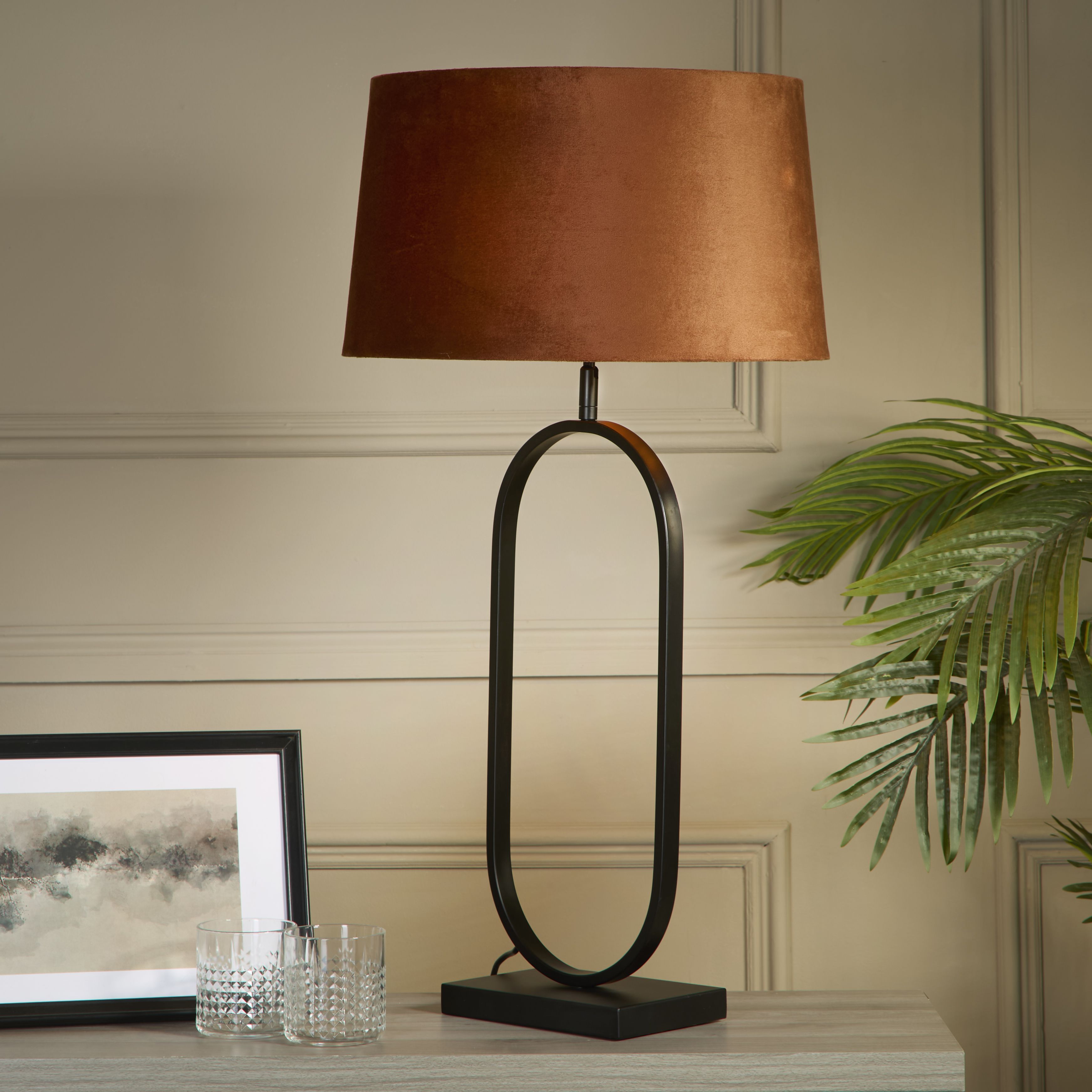 Modish table lamp with a burnt orange velvet shade styled on a light console table beside a framed print, two cut glass tumblers and a potted palm, shown switched on