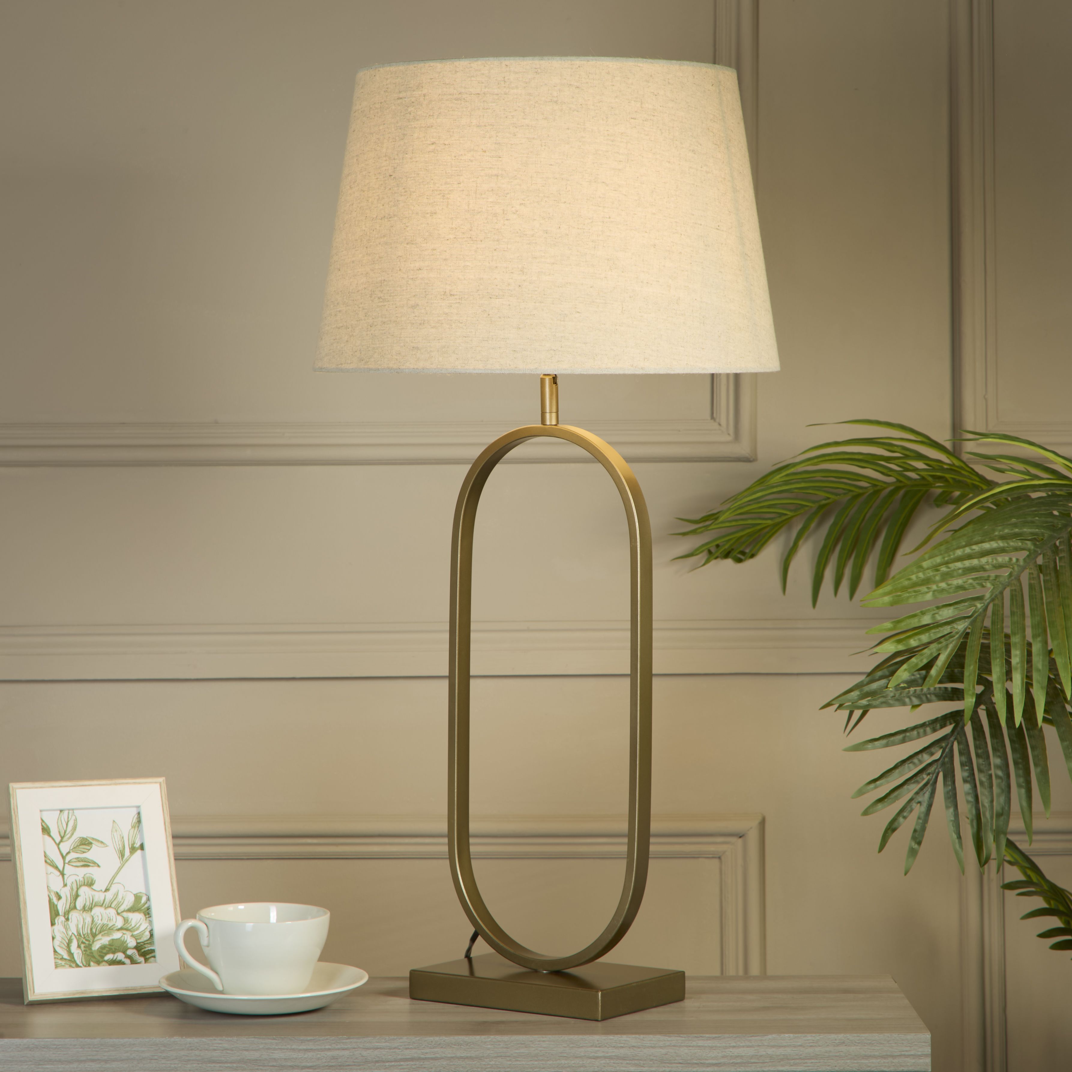 Bronze table lamp with a natural linen shade styled on a console table beside a teacup and saucer, a framed botanical print and a trailing palm plant, against a grey panelled wall