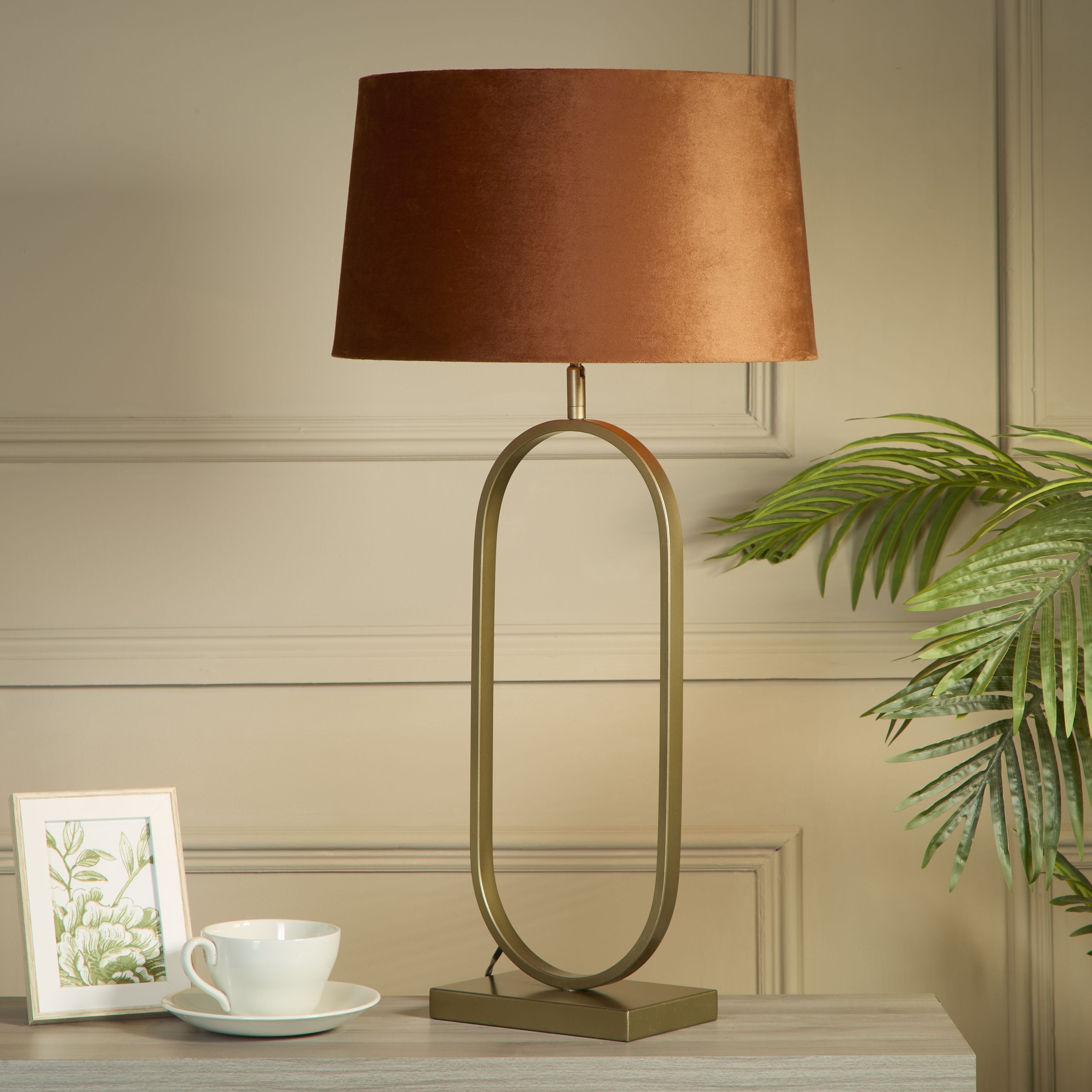 Bronze table lamp with a burnt orange velvet shade styled on a console table beside a teacup and saucer, a framed botanical print and a trailing palm plant, against a grey panelled wall