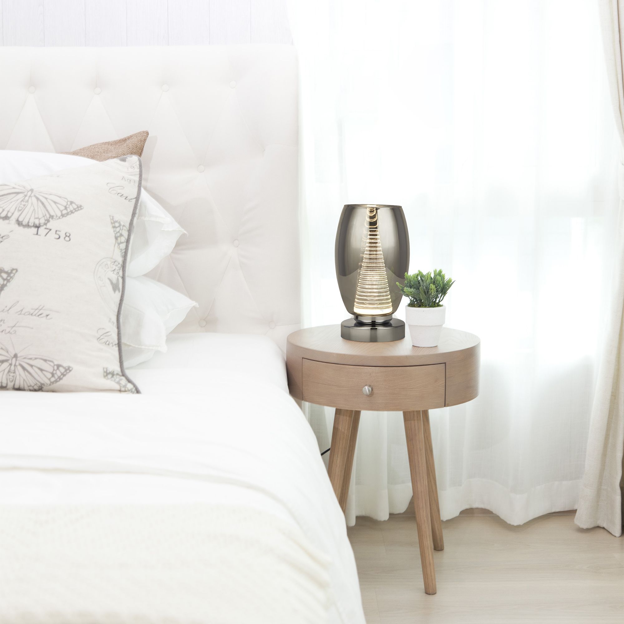 Searchlight Cyclone table lamp in chrome and smoked glass styled on a round bedside table next to a potted plant in a white bedroom