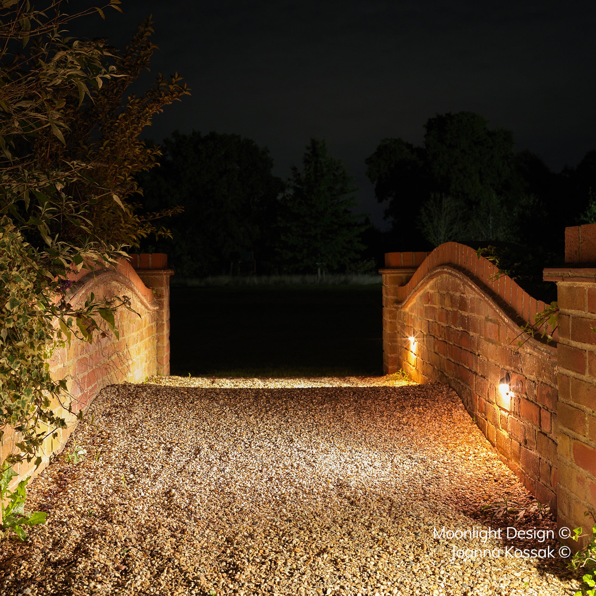 A gravel driveway between brick entrance piers lit by recessed Hunza Euro eyelid lights set into the brickwork at night. A Moonlight Design installation, not stock imagery.