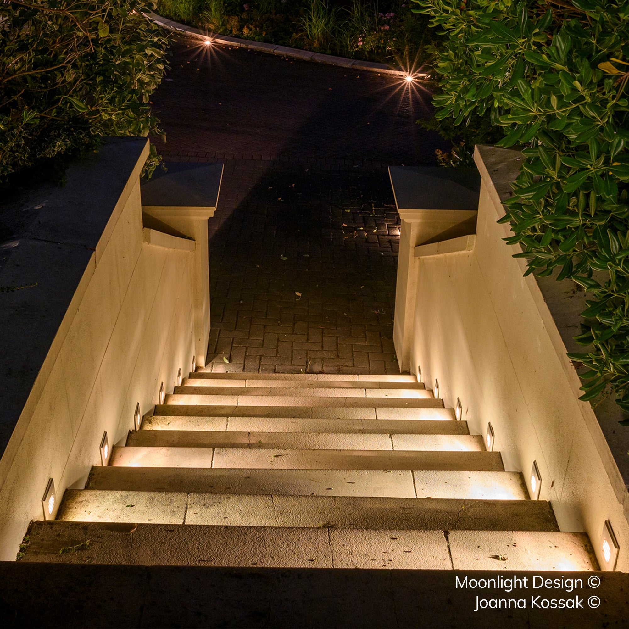 A wide stone staircase descending between two rendered walls to a brick courtyard at night, lit by rows of recessed louvred step lights along both sides. A Moonlight Design installation, not stock imagery.
