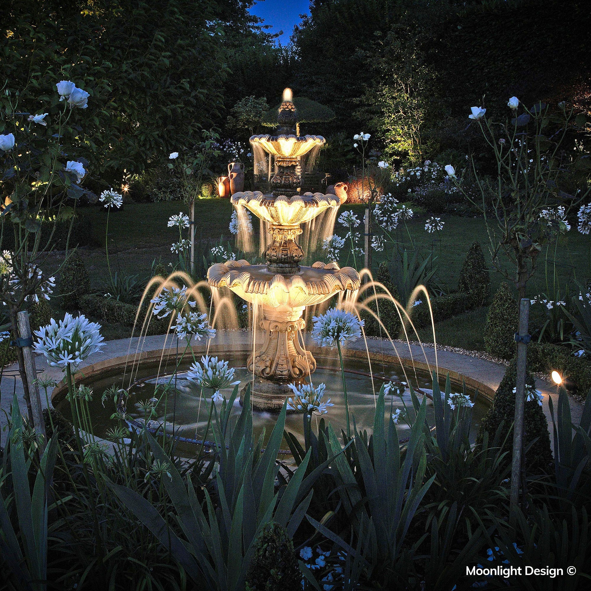 Warm Hunza pond lights illuminate the fountain and water features, adding depth and atmosphere to the traditional garden setting at dusk. A completed Moonlight Design pond lighting project.
