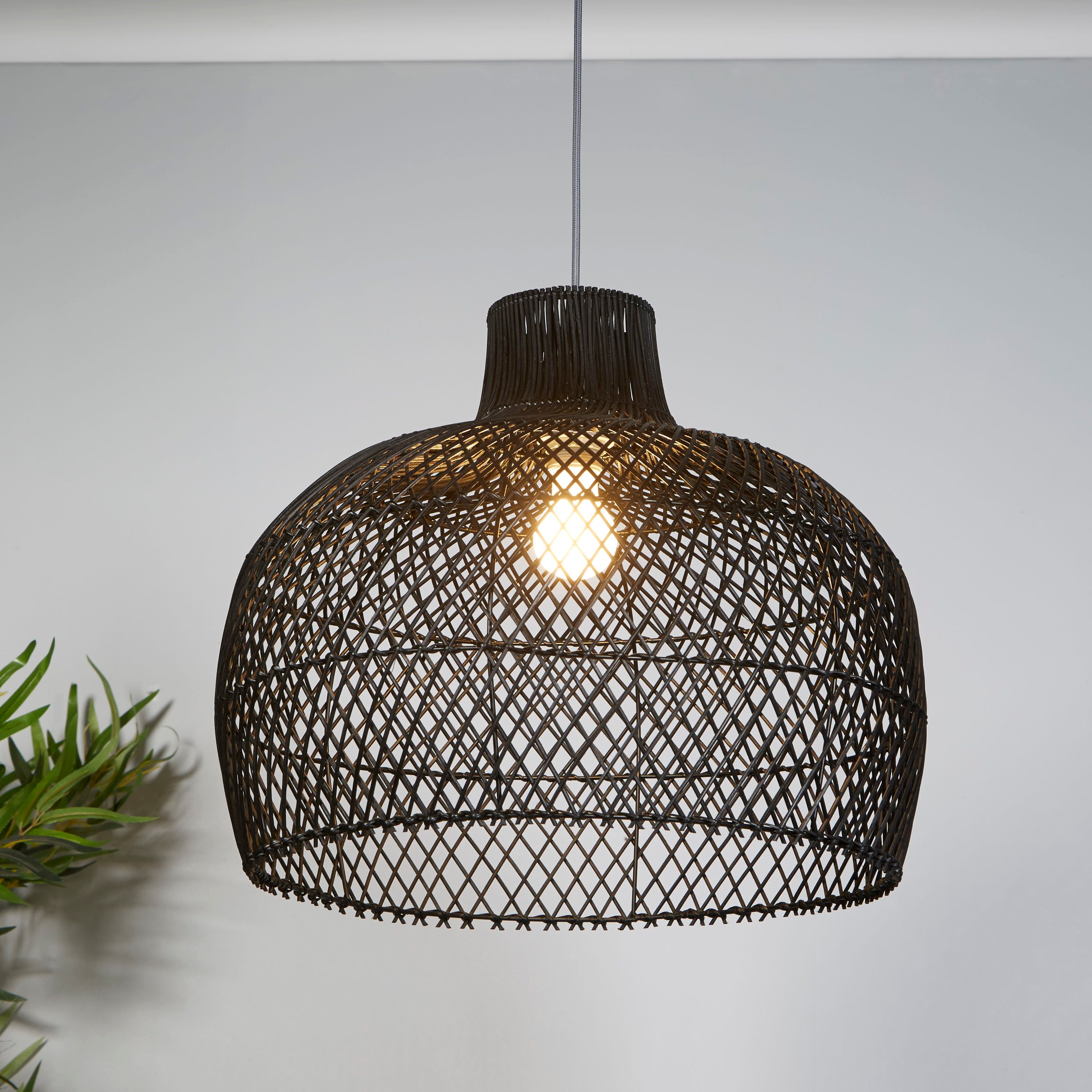 Black rattan bell pendant shade hung in a room with a grey wall and a potted plant, bulb lit