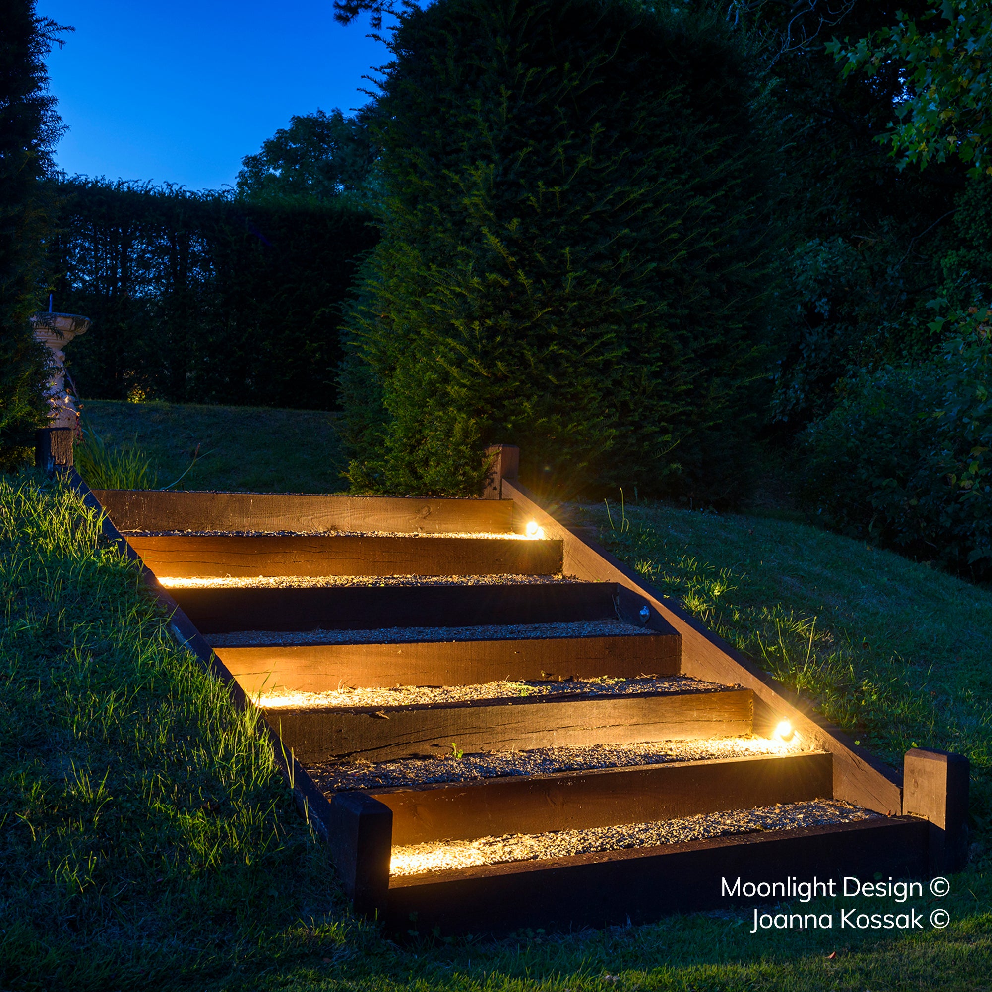A timber railway sleeper staircase descending through a garden at dusk, lit by recessed Hunza eyelid step lights along each riser. A Moonlight Design installation, not stock imagery.