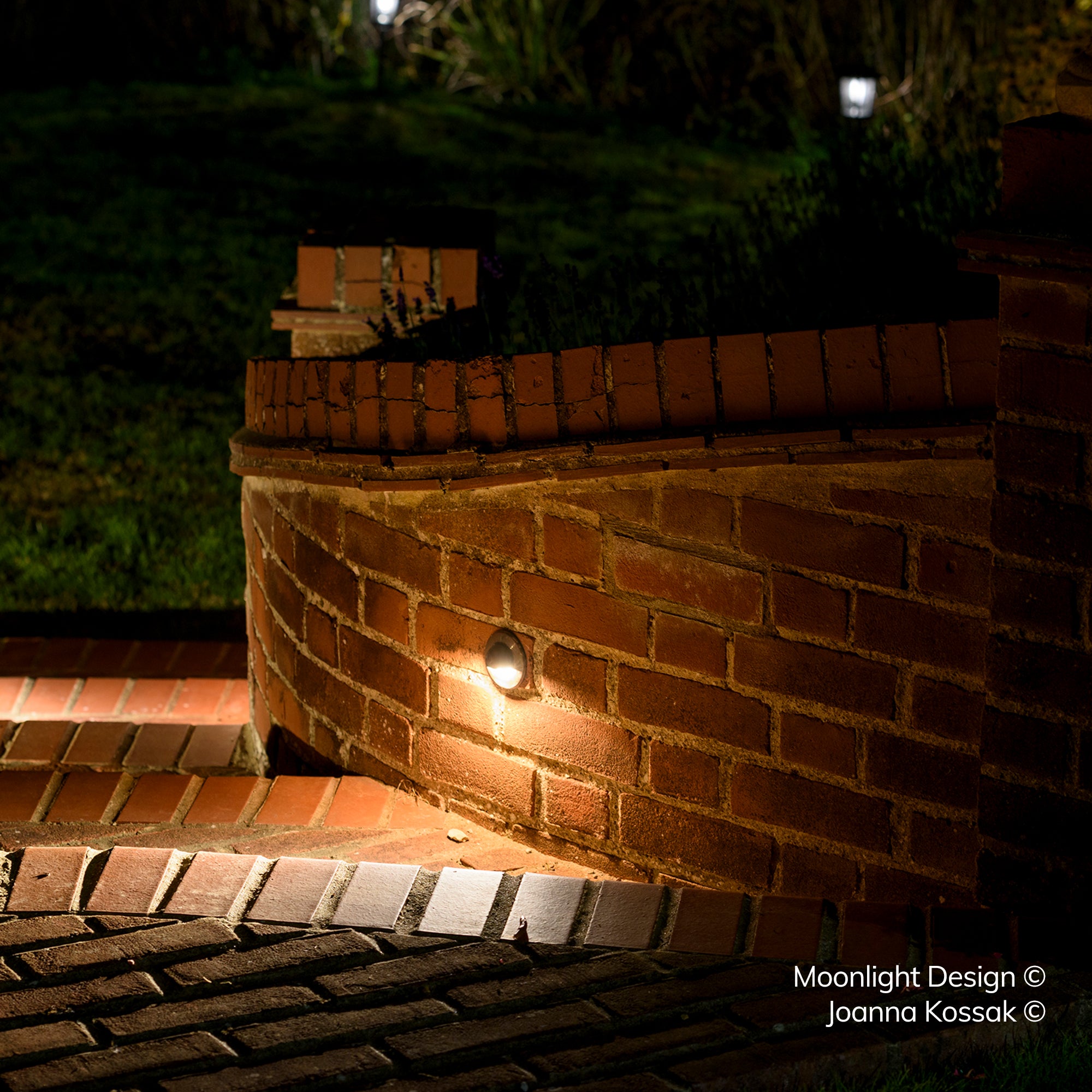 Close up of a Hunza eyelid step light recessed into a curved brick wall at the base of a flight of steps, lighting the brick paving below at night. A Moonlight Design installation, not stock imagery.
