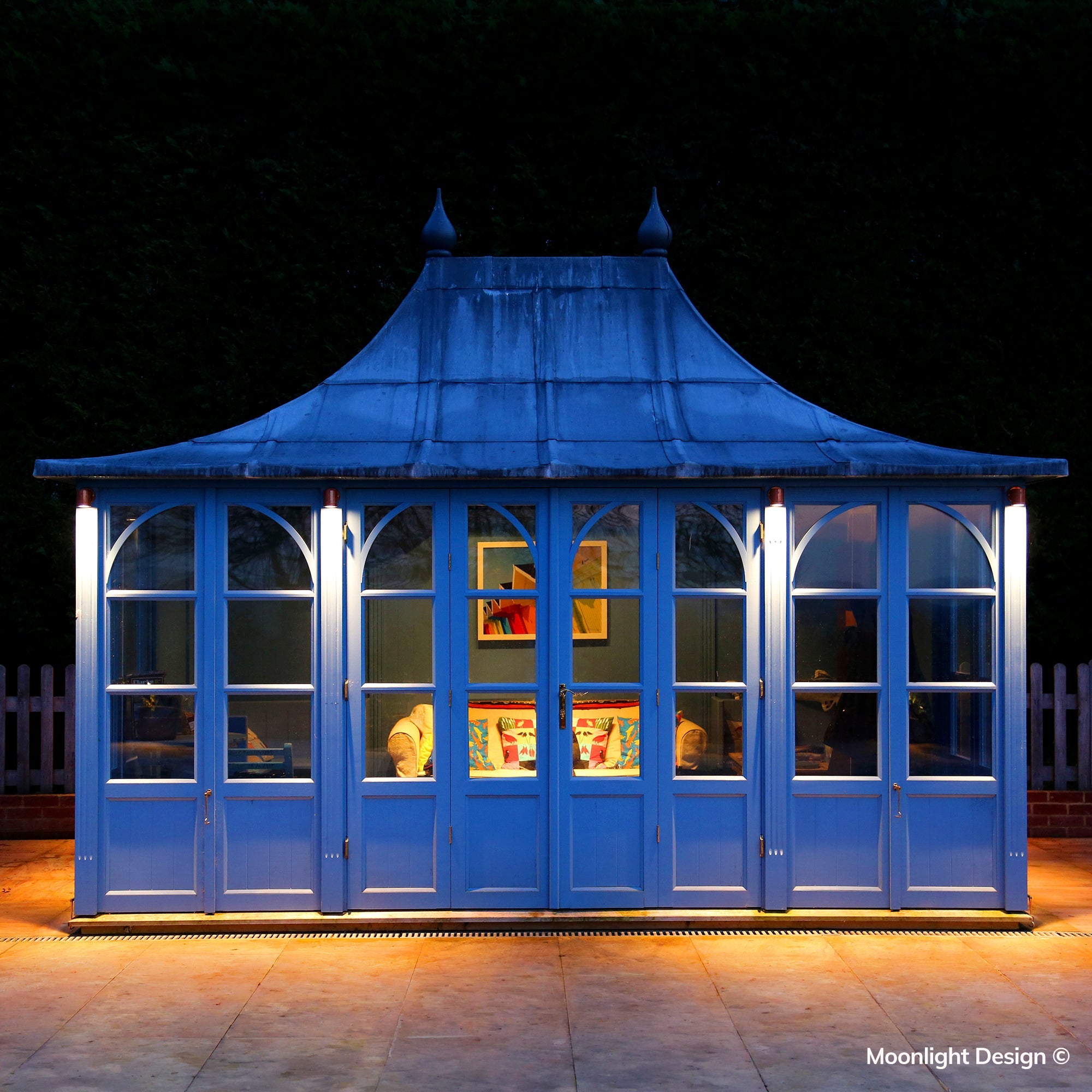 Moonlight Design lifestyle photograph of Hunza wall down lights washing light down the corner posts of a blue-painted garden pavilion at dusk.