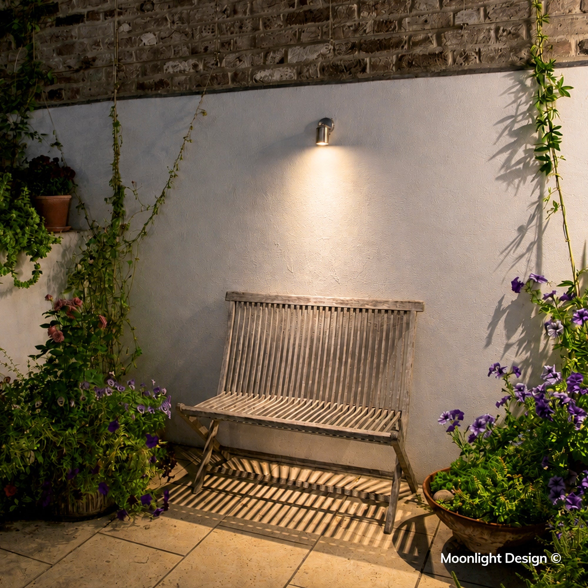 Moonlight Design lifestyle photograph of a Hunza wall down light above a weathered wooden bench, surrounded by climbing plants and flowering containers at night.