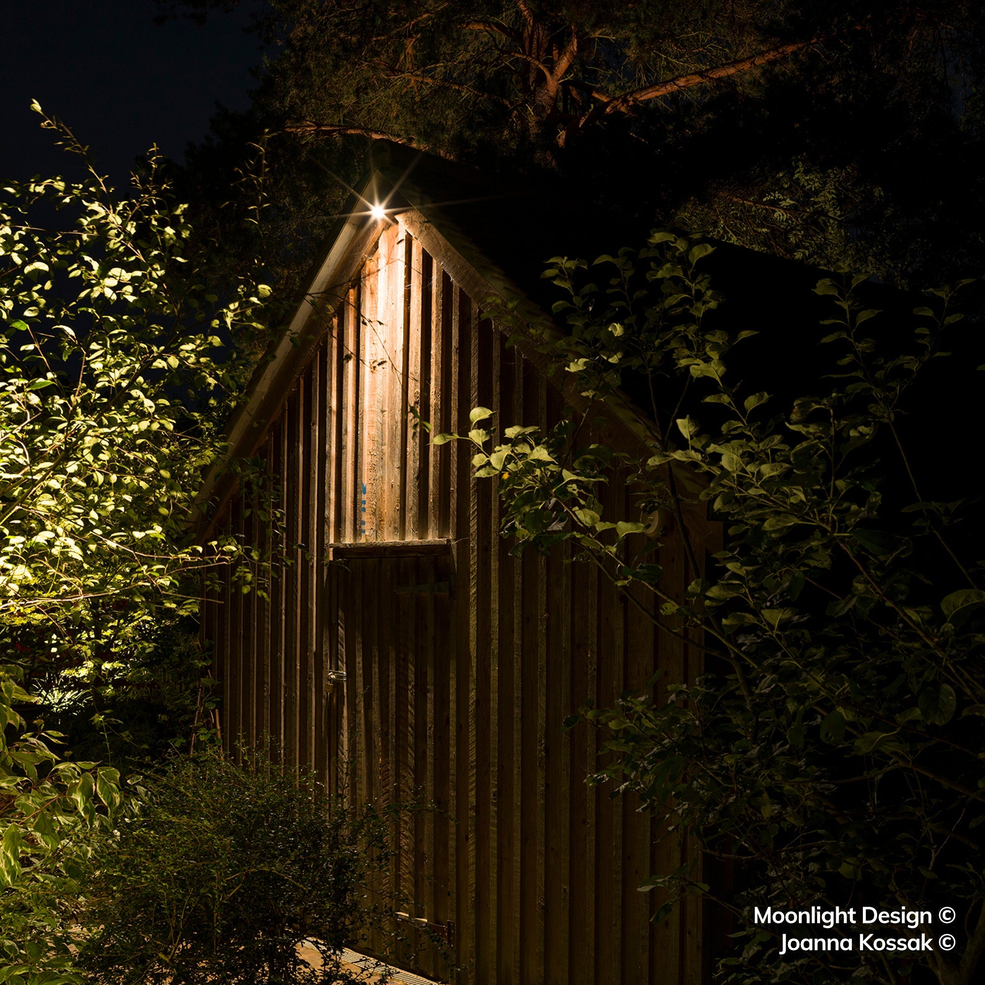 Moonlight Design lifestyle photograph of a Hunza wall down light fixed to the gable apex of a timber-clad garden building, lighting the boarded end at night.