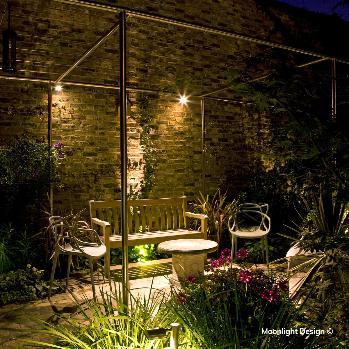 Moonlight Design lifestyle photograph of a Hunza wall spot lighting a brick wall and birdhouse above a wooden bench in a pergola-covered garden seating area at night.