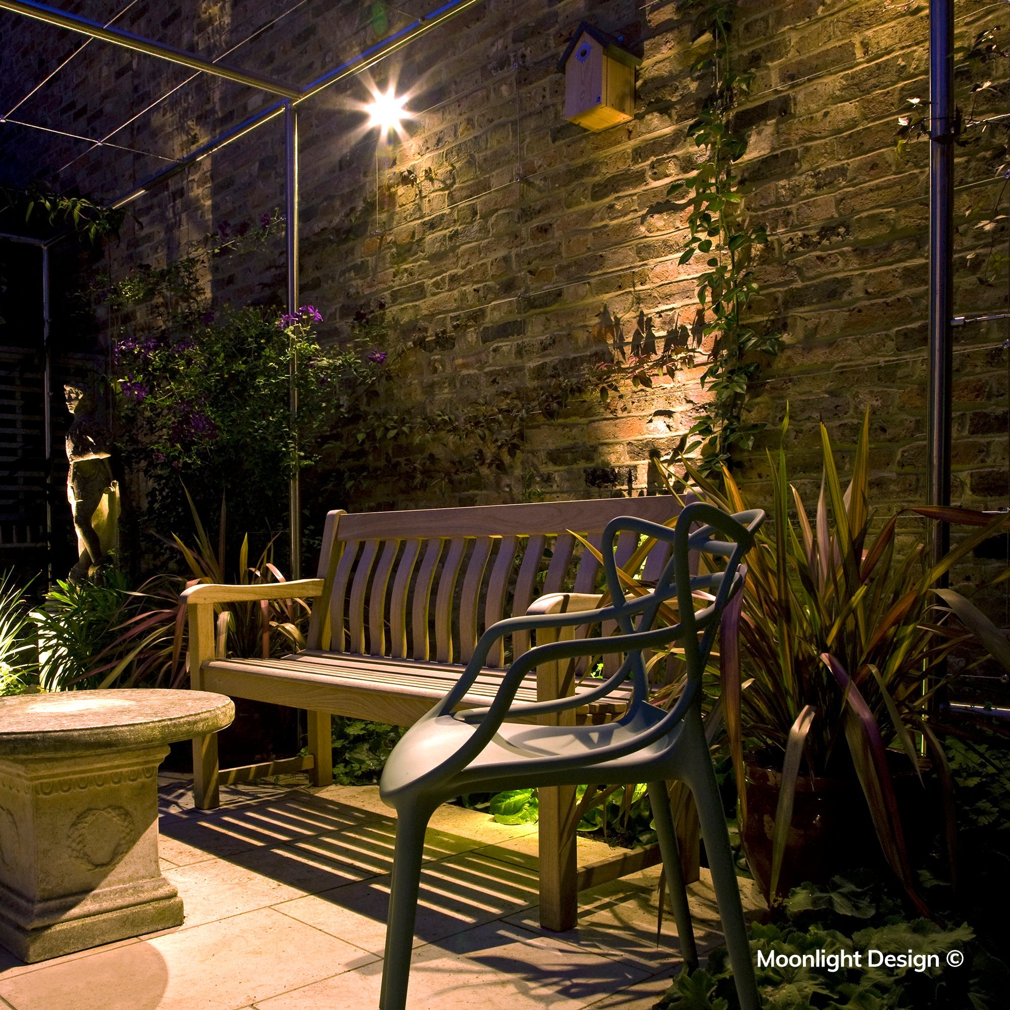 Moonlight Design lifestyle photograph of Hunza wall spots lighting a brick wall and birdhouse above a garden bench, framed by magenta flowering plants in a pergola seating area at night.