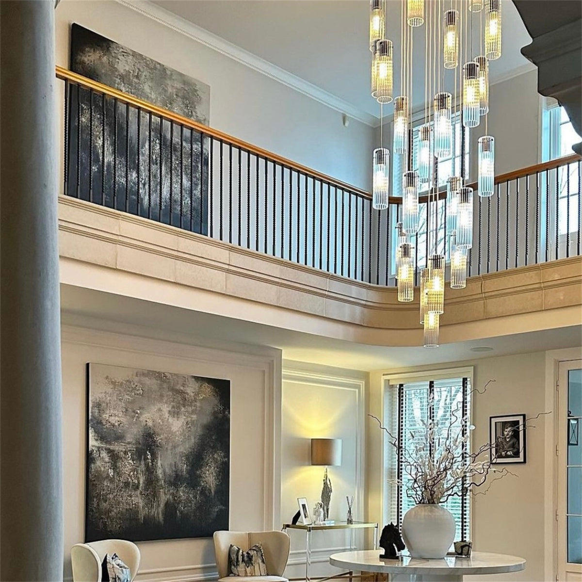 Many lit ribbed glass cylinder pendants with brass tops hanging at different heights in a two storey hall beside a curved landing balustrade, from a Leverint cascade installation
