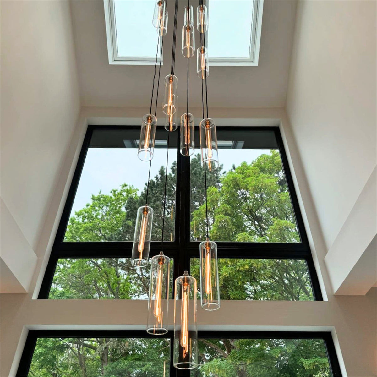 Tall clear glass cylinder pendants with long filament bulbs hanging at different heights in front of a large double height window, from a Leverint cascade installation