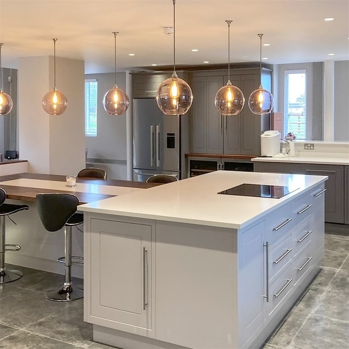 Line of round clear glass pendants with lit filament bulbs hanging above a white kitchen island and breakfast bar in a grey painted kitchen