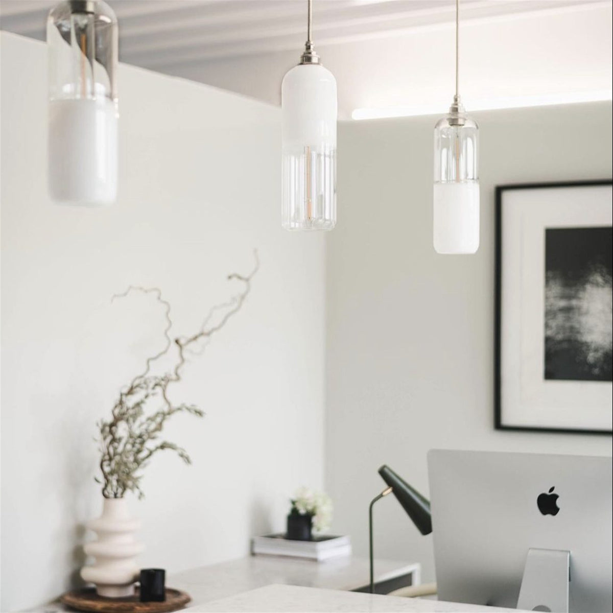 Three capsule shaped pendants in clear and white glass hanging at different heights above a desk in a bright white room