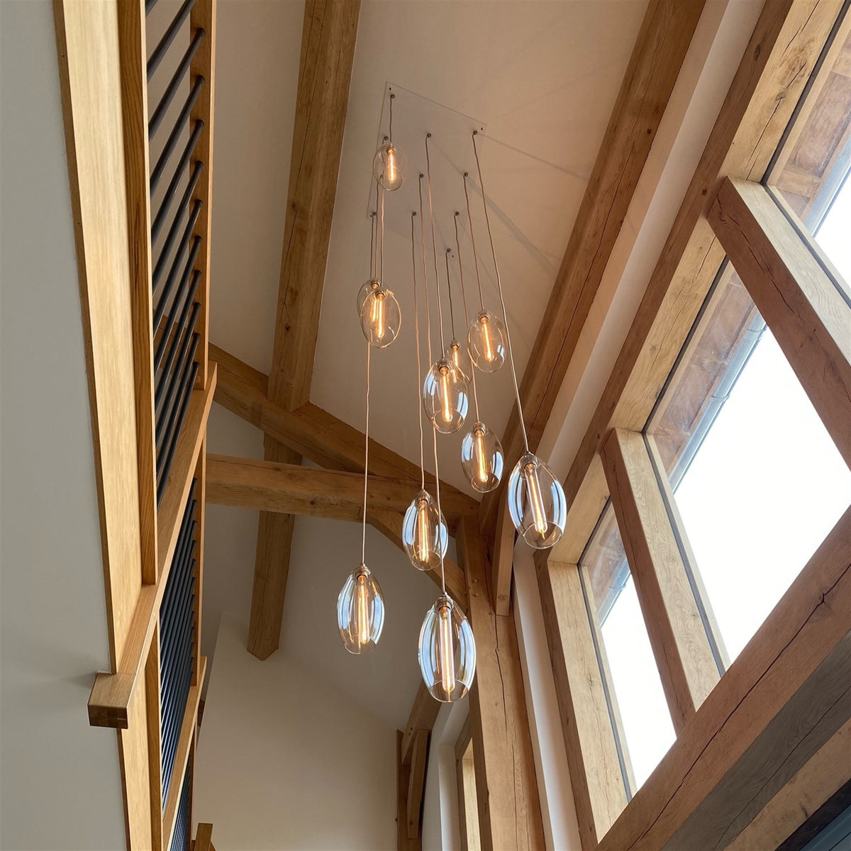 Group of teardrop shaped glass pendants with lit filament bulbs hanging at different lengths from a rectangular ceiling plate in a double height space with exposed oak beams and tall windows