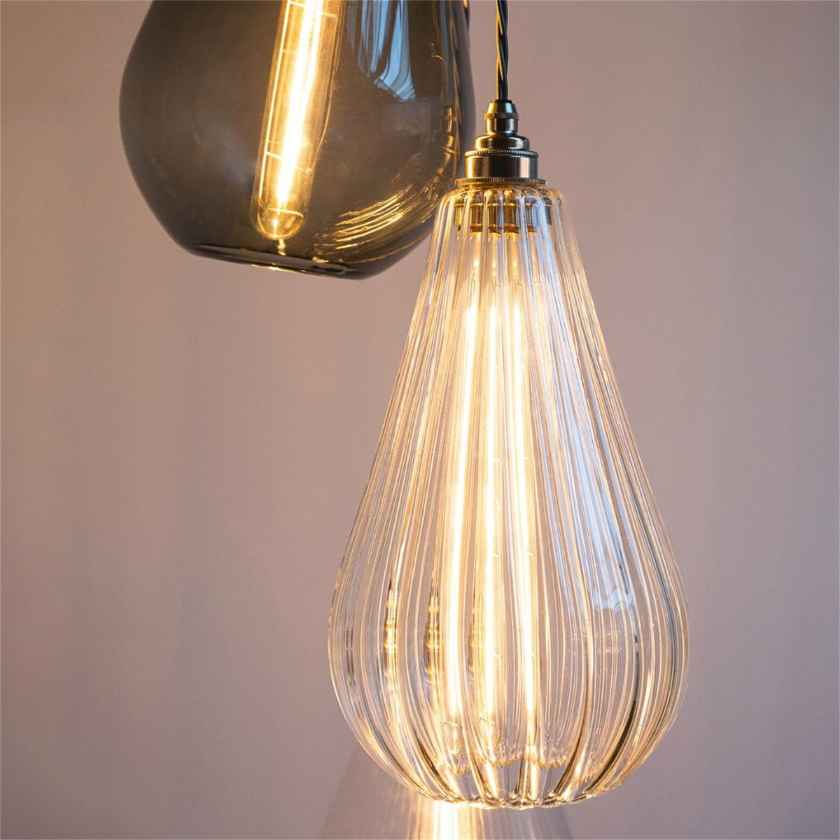 Close up of a lit clear vertically ribbed teardrop glass pendant with a metal lamp holder and twisted flex, with a smoky grey glass pendant above it