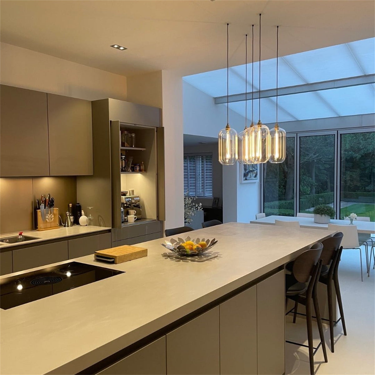 Cluster of lit Leverint Charlton pendants in glass with brass toned fittings, hung at different heights over a kitchen island