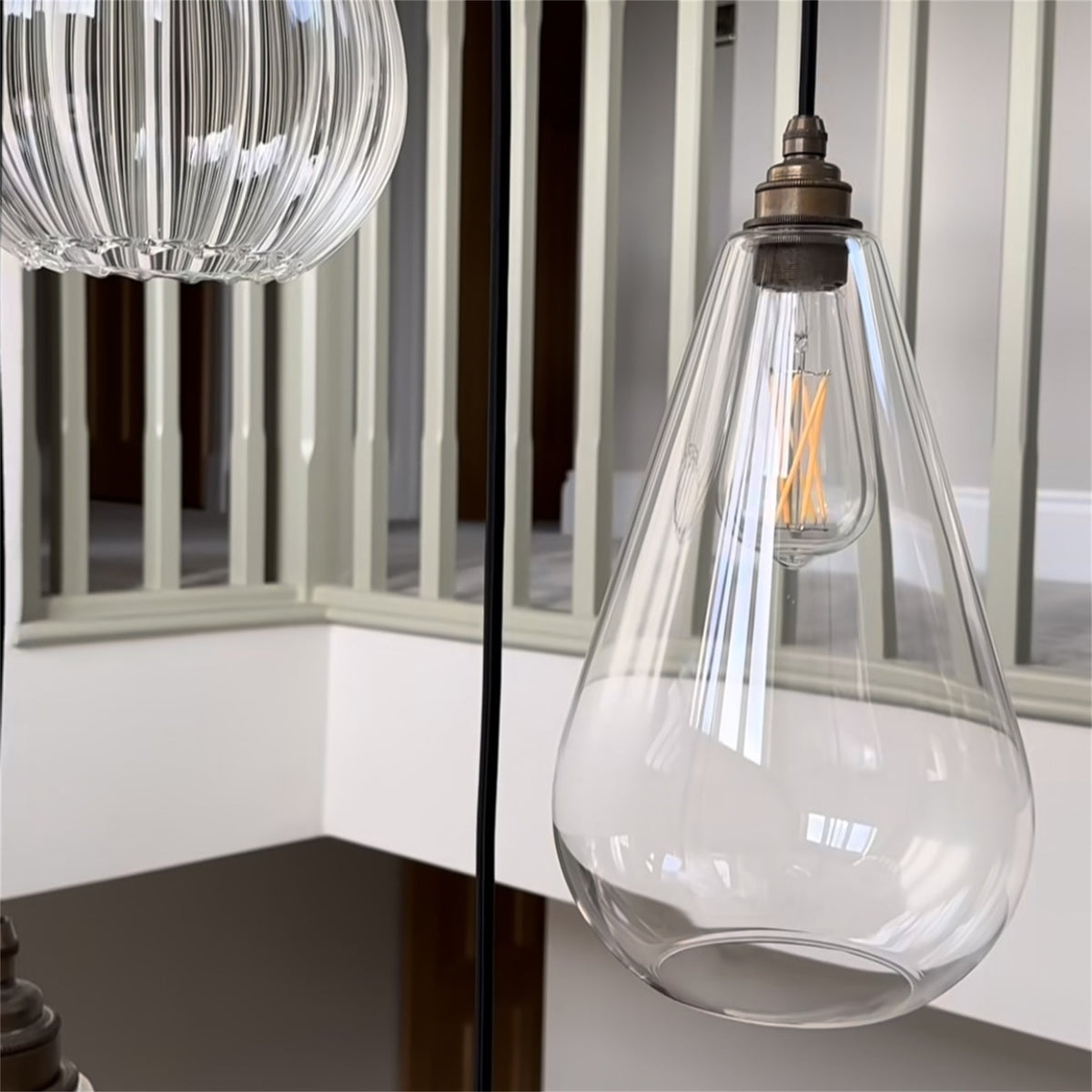 Close up of a clear glass Leverint Kingston teardrop pendant with a bronze toned fitting and lit filament bulb, with a ribbed glass shade beside it
