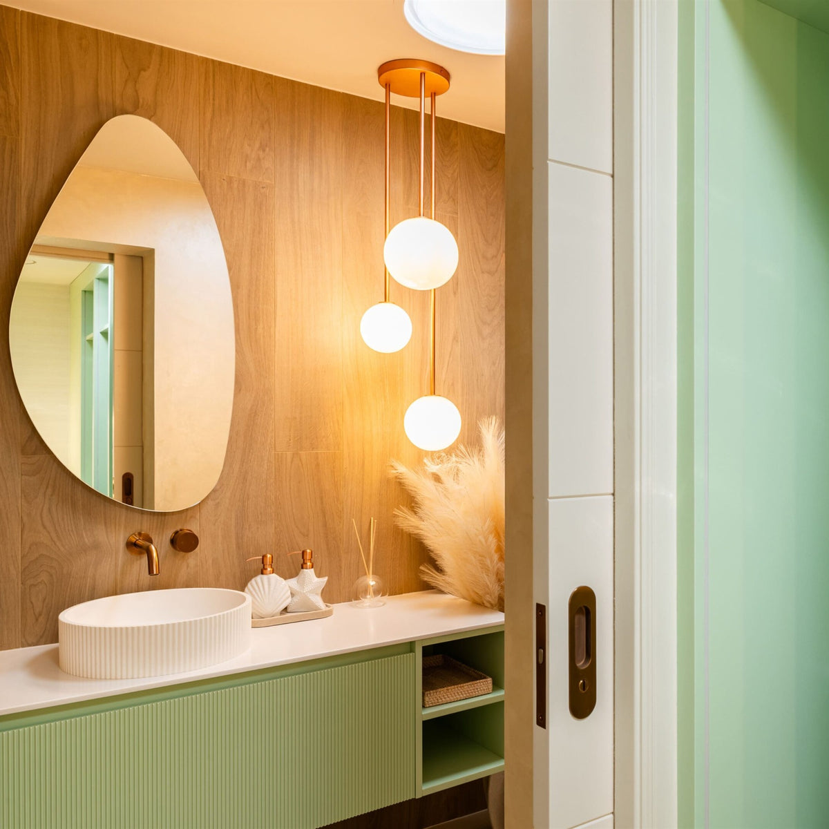 Three opal white glass globes in graduated sizes lit warm on copper coloured rods from a round copper ceiling plate, beside an organic shaped mirror and a wood effect wall