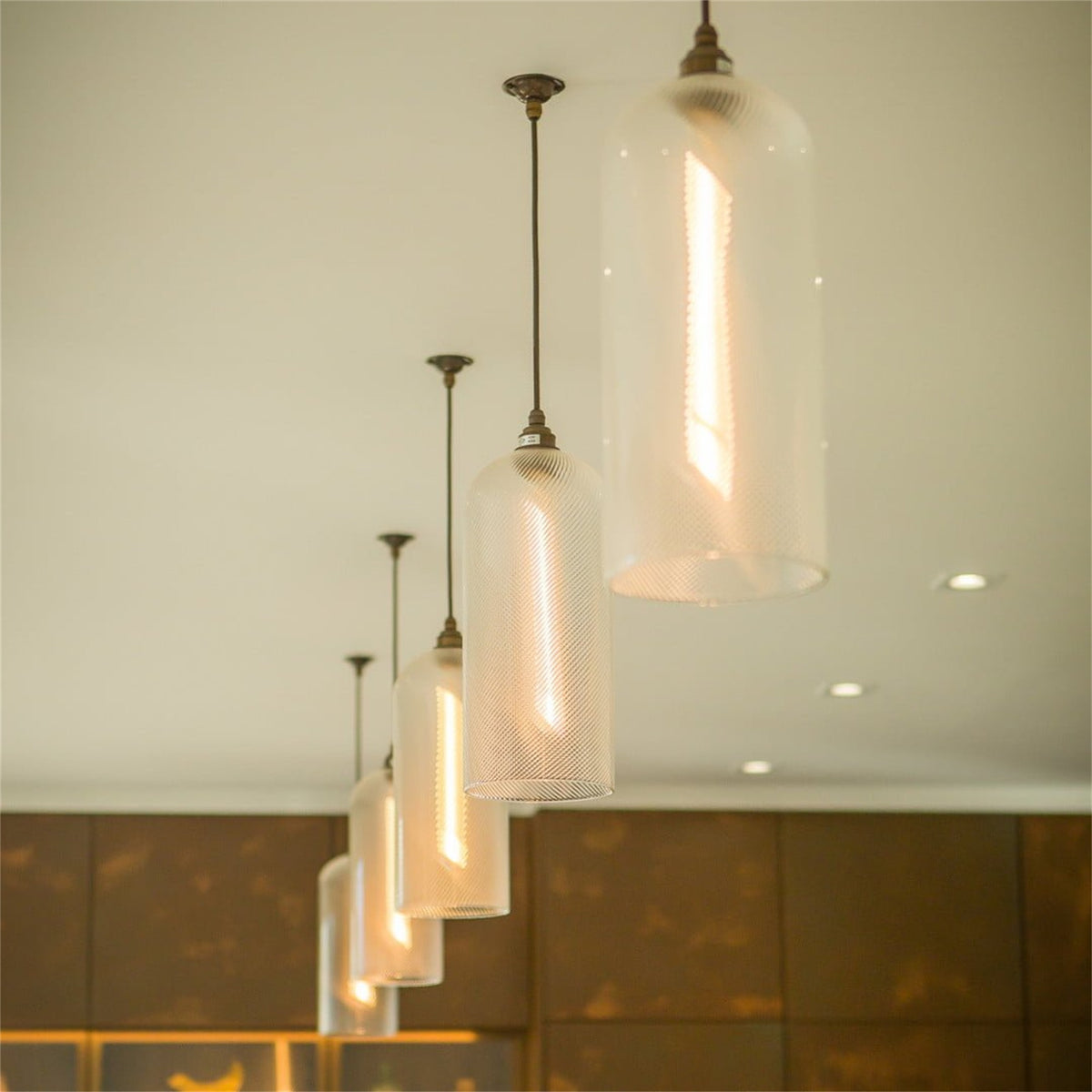 Row of lit Leverint Richmond pendants in finely ribbed glass with bronze toned fittings, hung in a line at descending heights from a ceiling