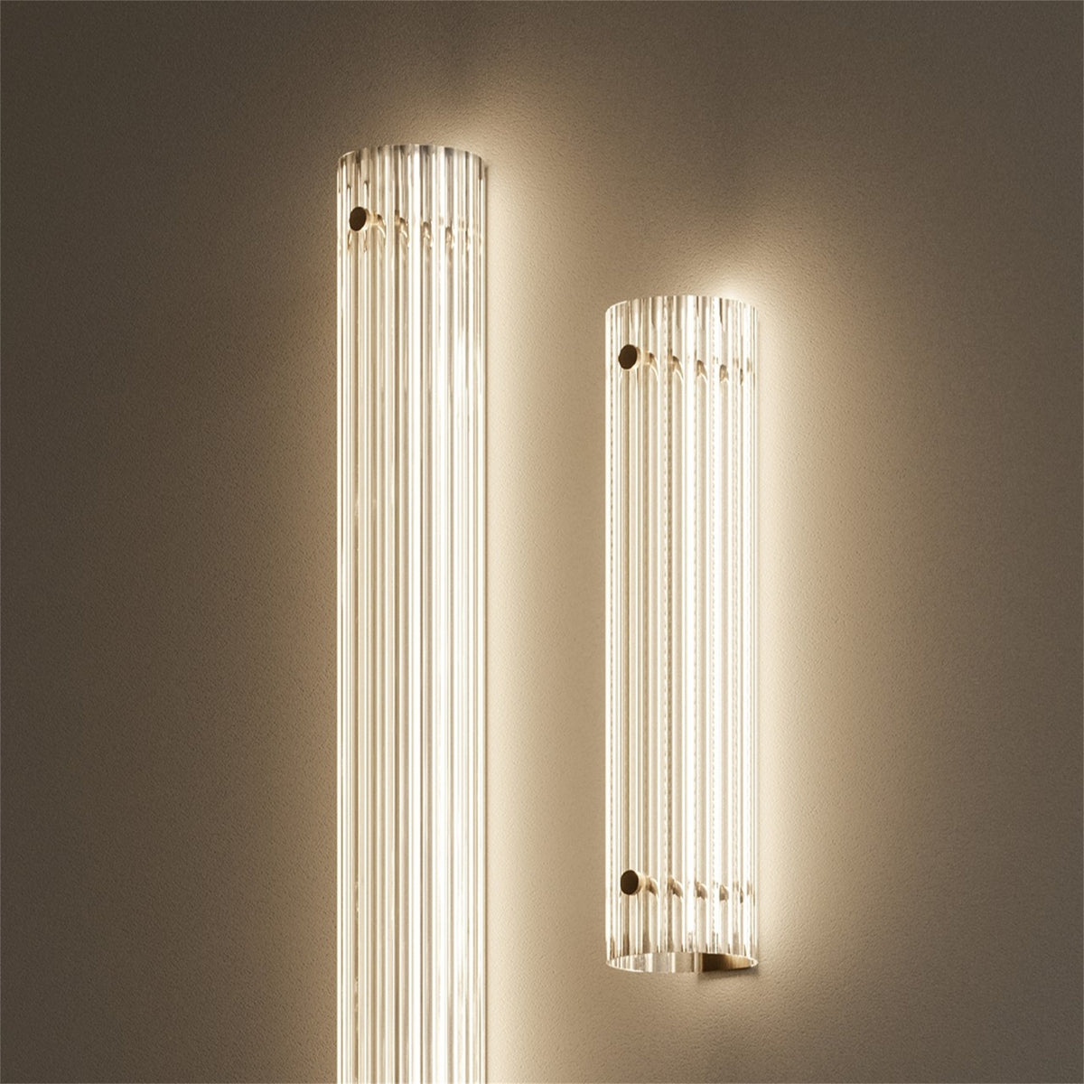 Two lit Leverint Temple Pillar Lights of different heights on a warm taupe wall, ribbed glass columns glowing, close up