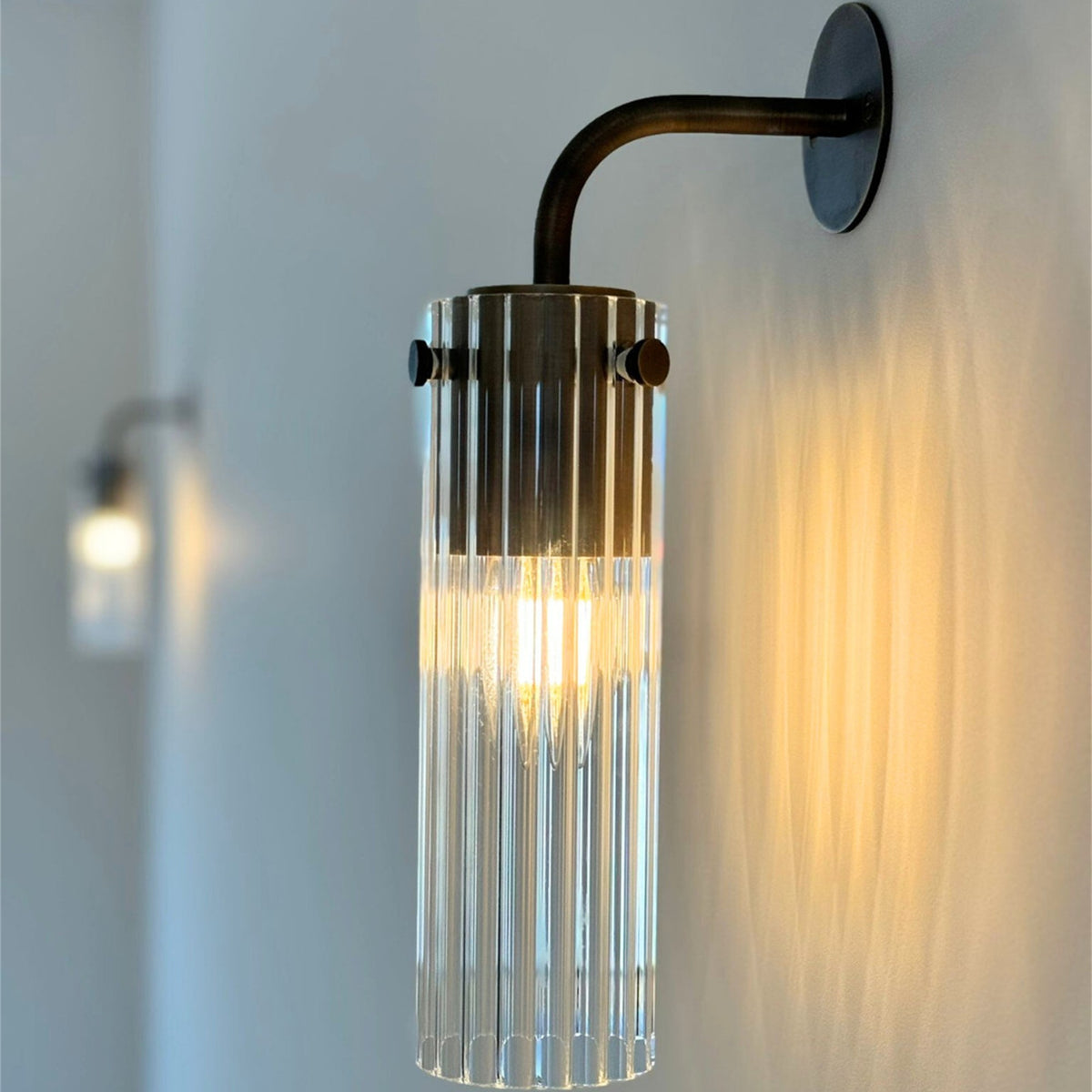 Leverint Temple Wall Light installed on a pale wall and lit, a ribbed glass cylinder with a metal collar on a curved dark metal arm, with a second light blurred behind