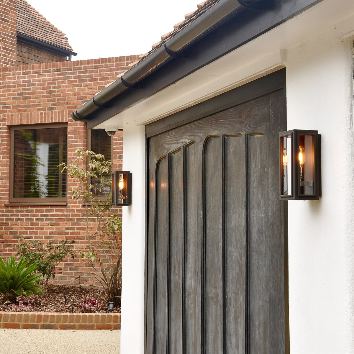 Two lit Leverint box wall lanterns with dark metal frames and glass panels on a white wall beside a dark panelled door and a brick building