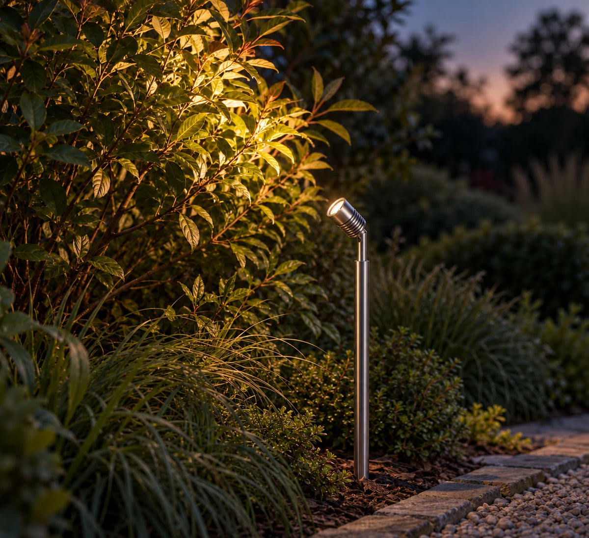 LuxR stainless steel pole spot in a planted garden border at dusk, lighting a leafy shrub