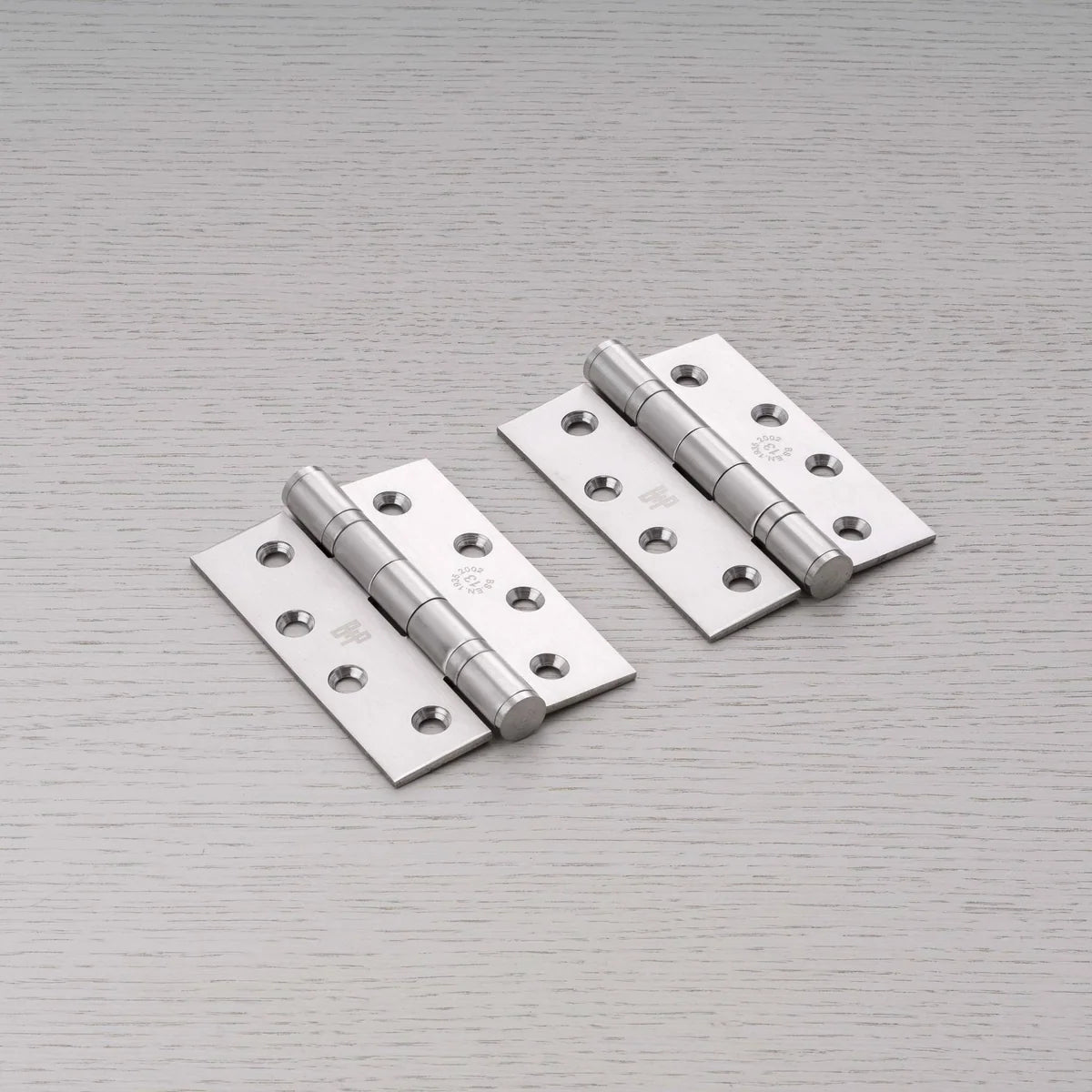 Pair of Buster + Punch steel door hinges laid on a pale grey wood grain surface, with countersunk screw holes