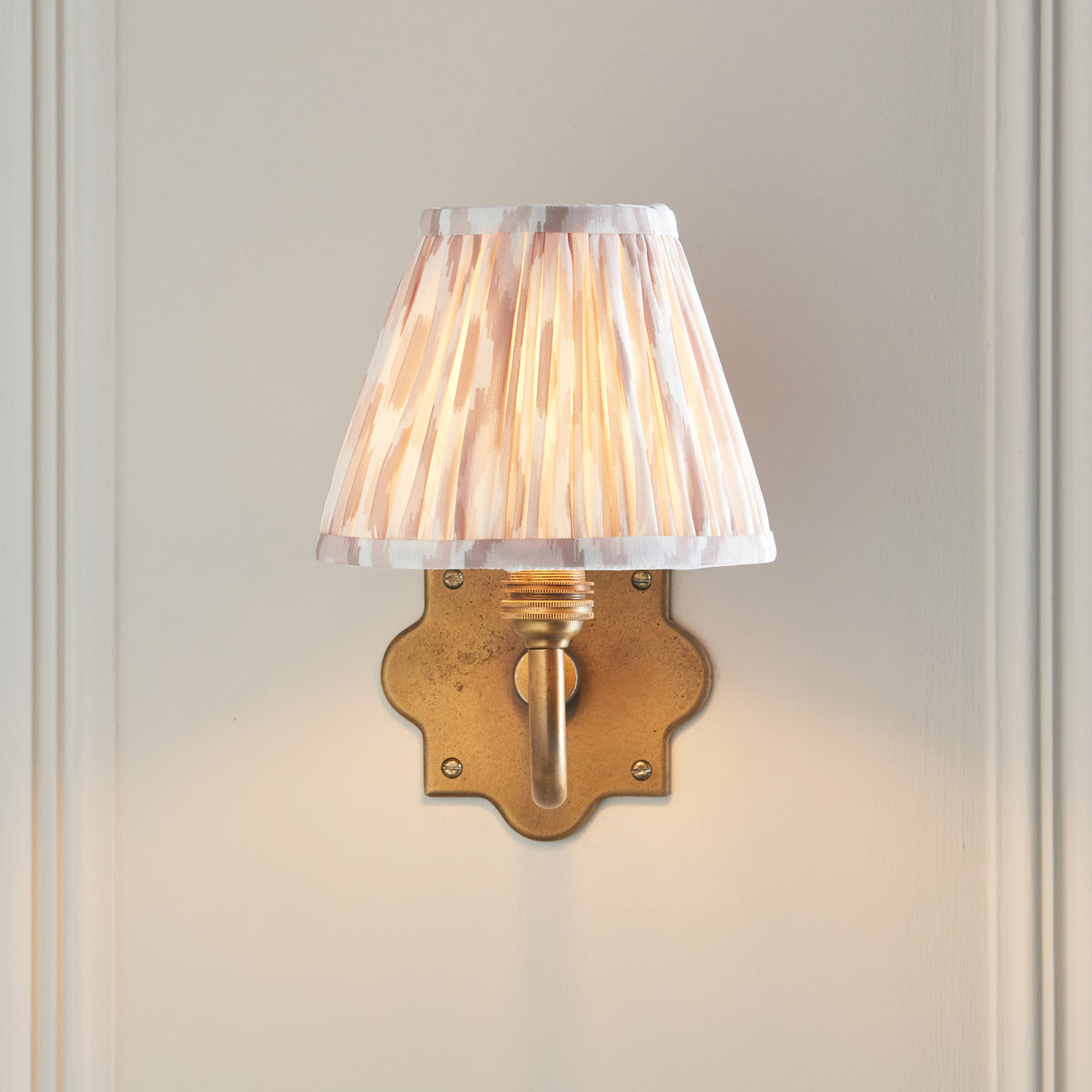 Endon Anvil Curve wall light lit on a cream panelled wall, the ikat shade sitting directly above the quatrefoil brass backplate