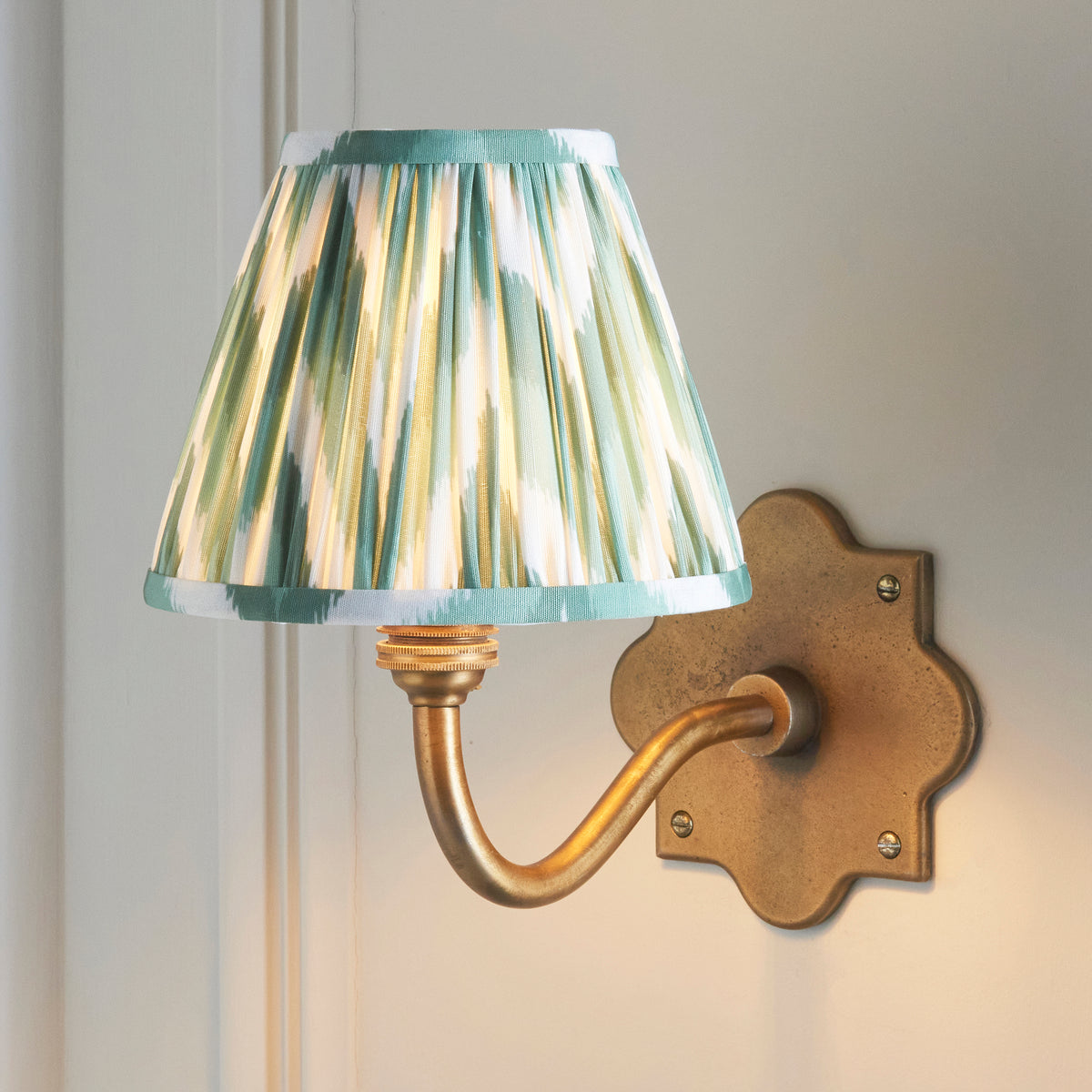 Endon Anvil Curve wall light lit on a pale grey panelled wall, close up of the green zigzag pleated shade and antique brass backplate