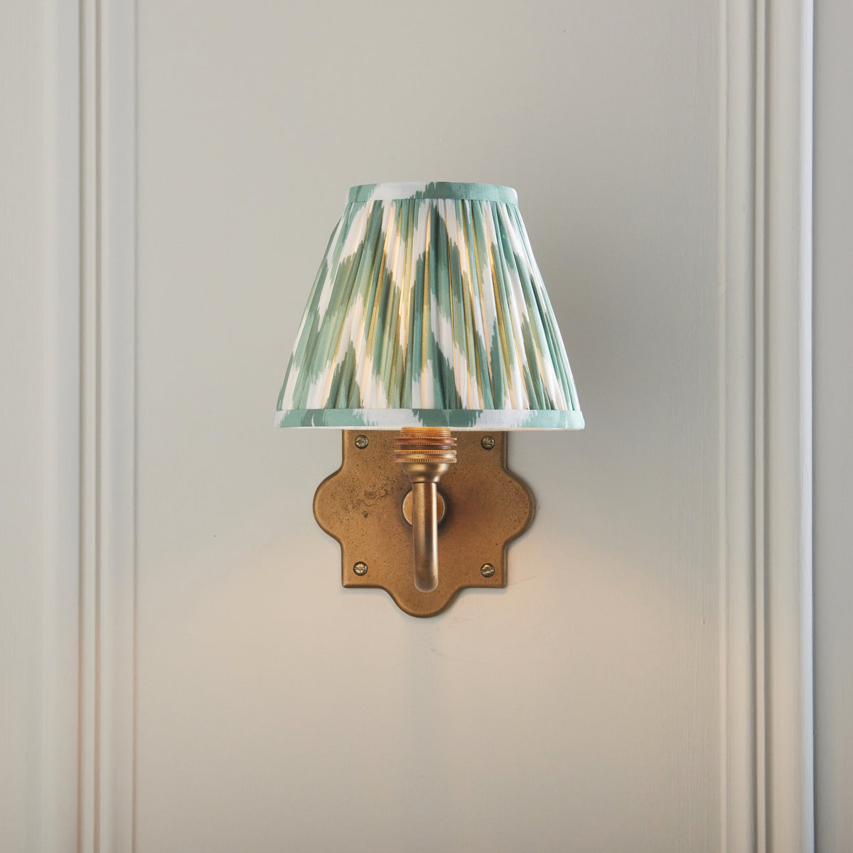 Endon Anvil Curve wall light lit on pale panelling, green zigzag shade sitting directly above the scalloped antique brass backplate